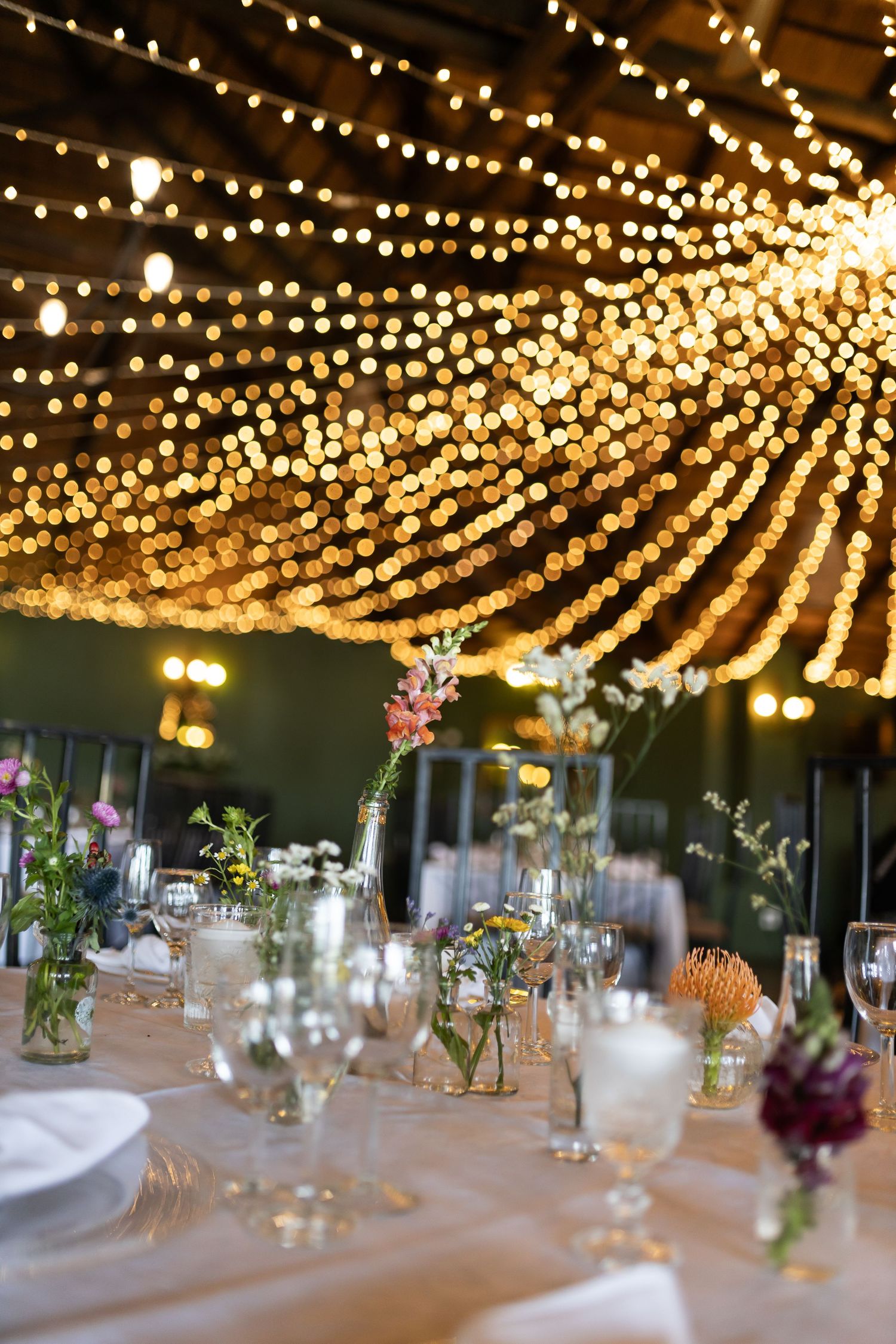 Elegant wedding reception table setting with string lights overhead and small floral centerpieces in glass vases.