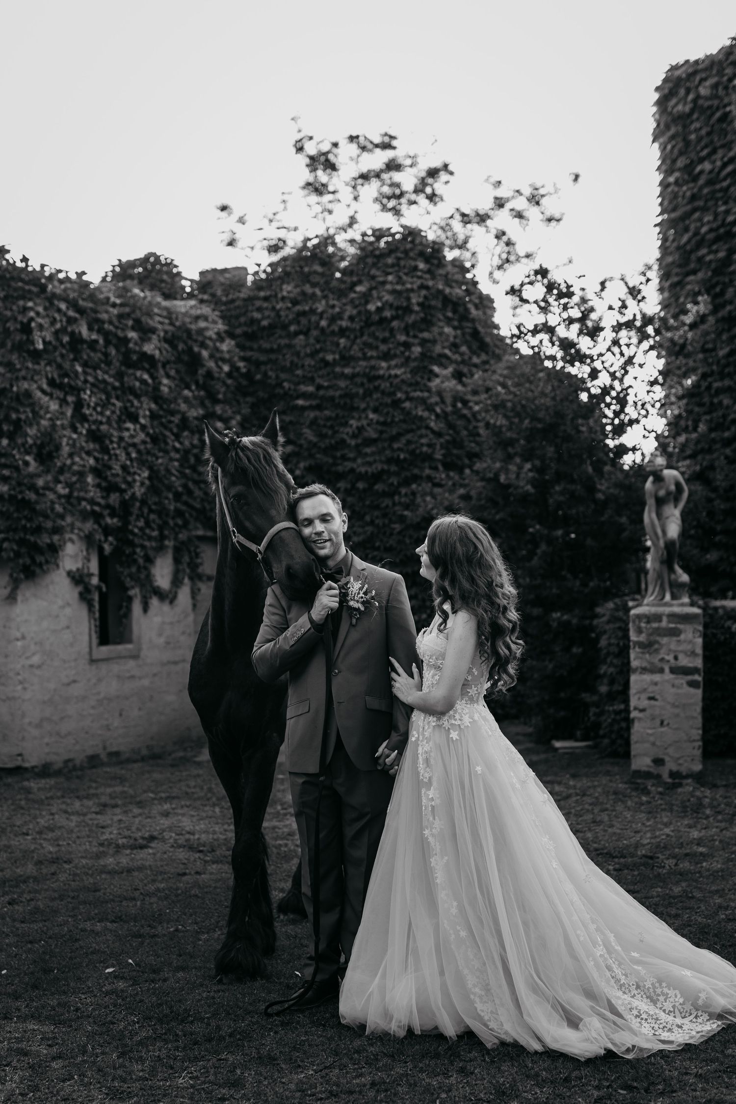 Black and white wedding photo with newlyweds and a horse against an ivy-covered castle wall.