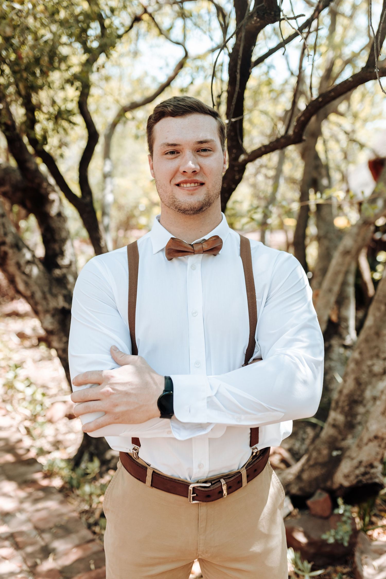 Groomsman in white shirt and brown suspenders crosses arms while posing in wooded area.