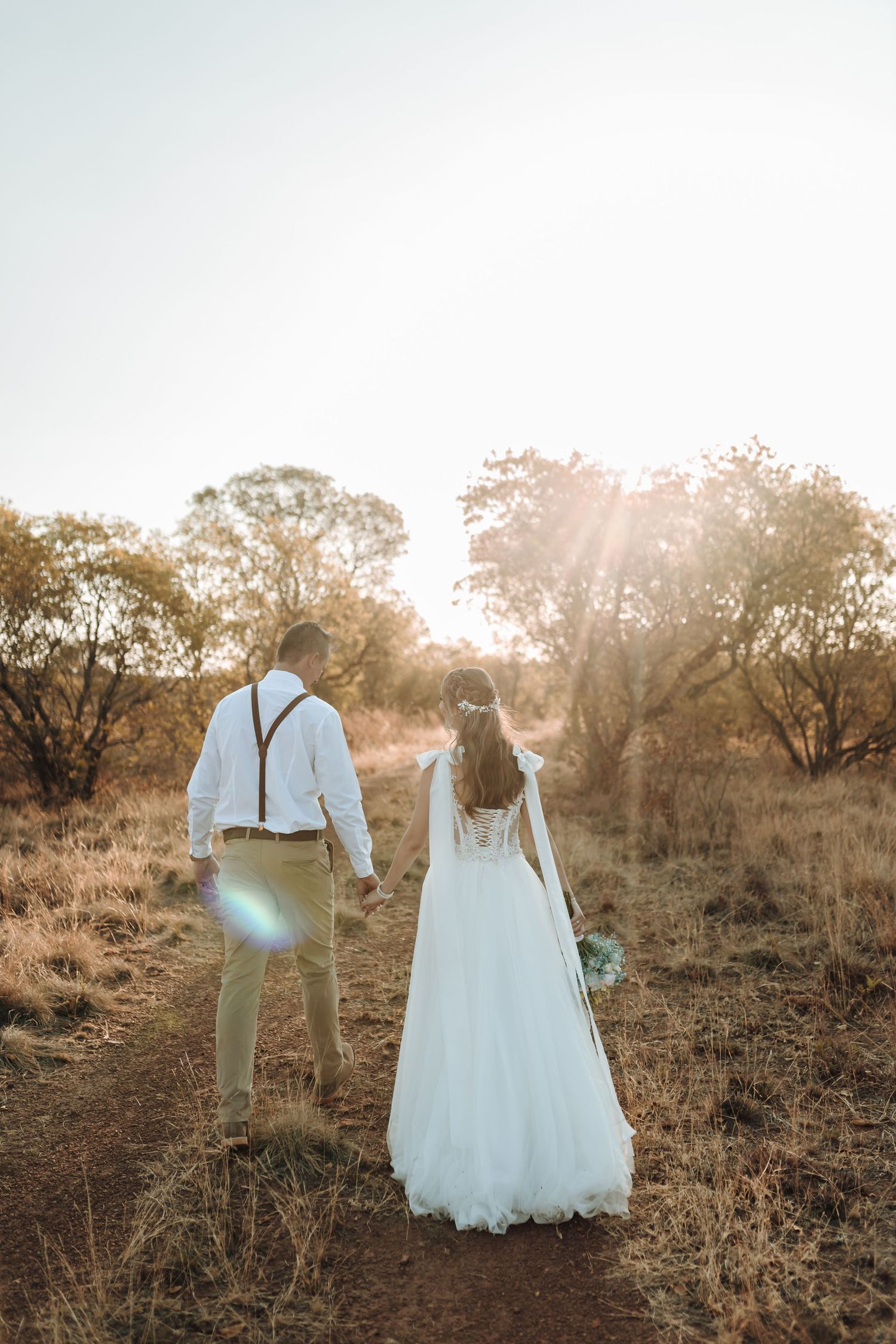Couple in wedding attire walks hand in hand through a sunlit outdoor path.