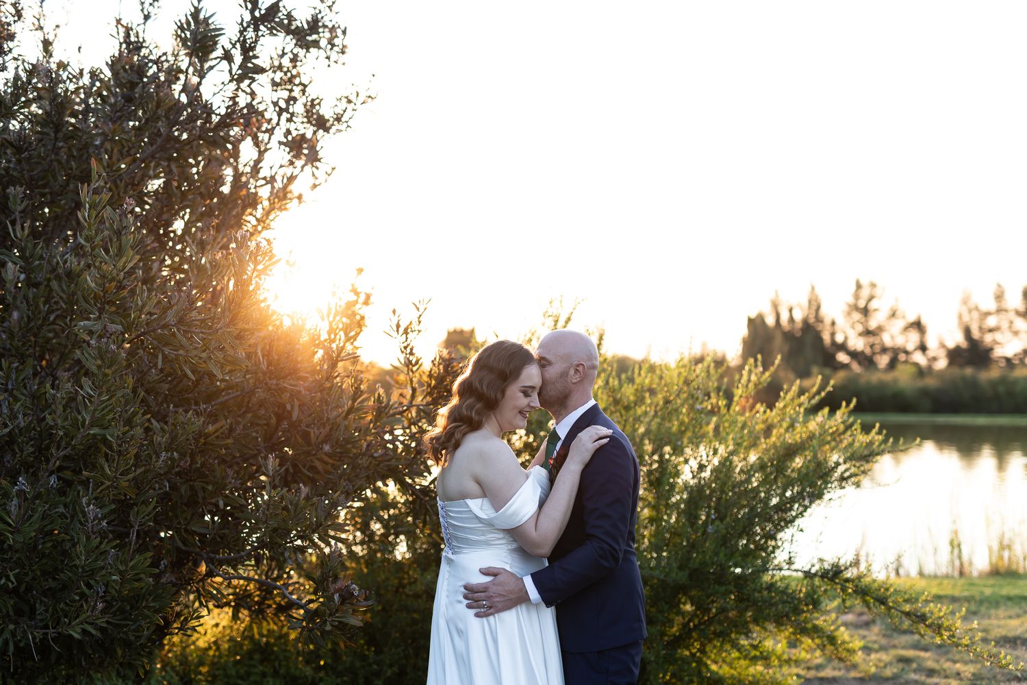 A couple in wedding attire embraces near water during golden hour sunset.