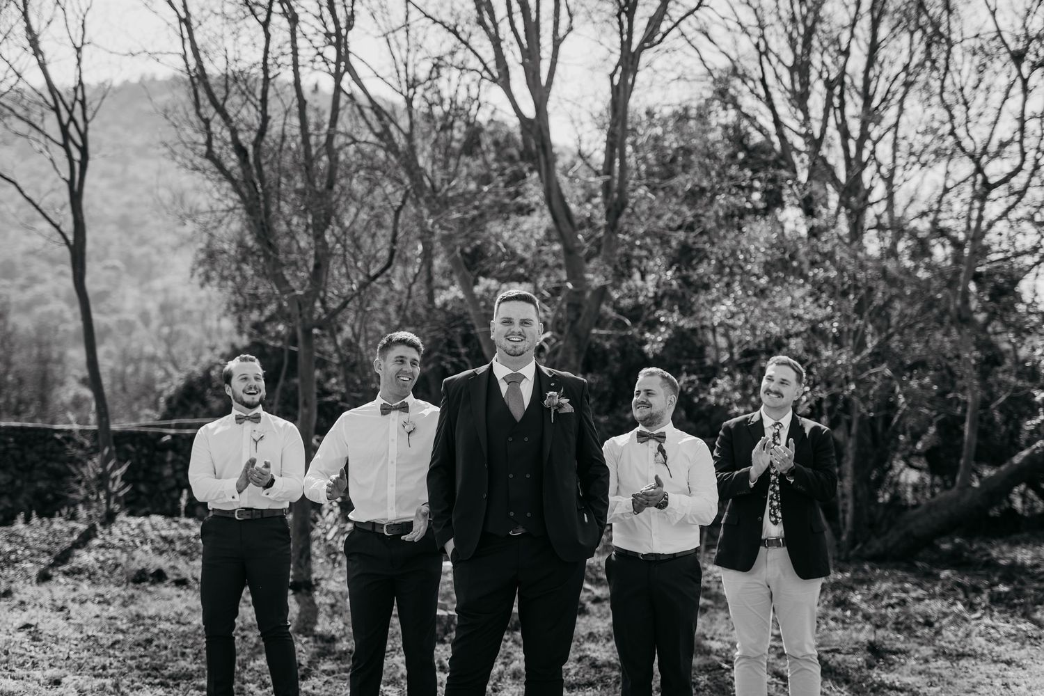 A black and white photograph of groomsmen standing together outdoors with trees in the background.