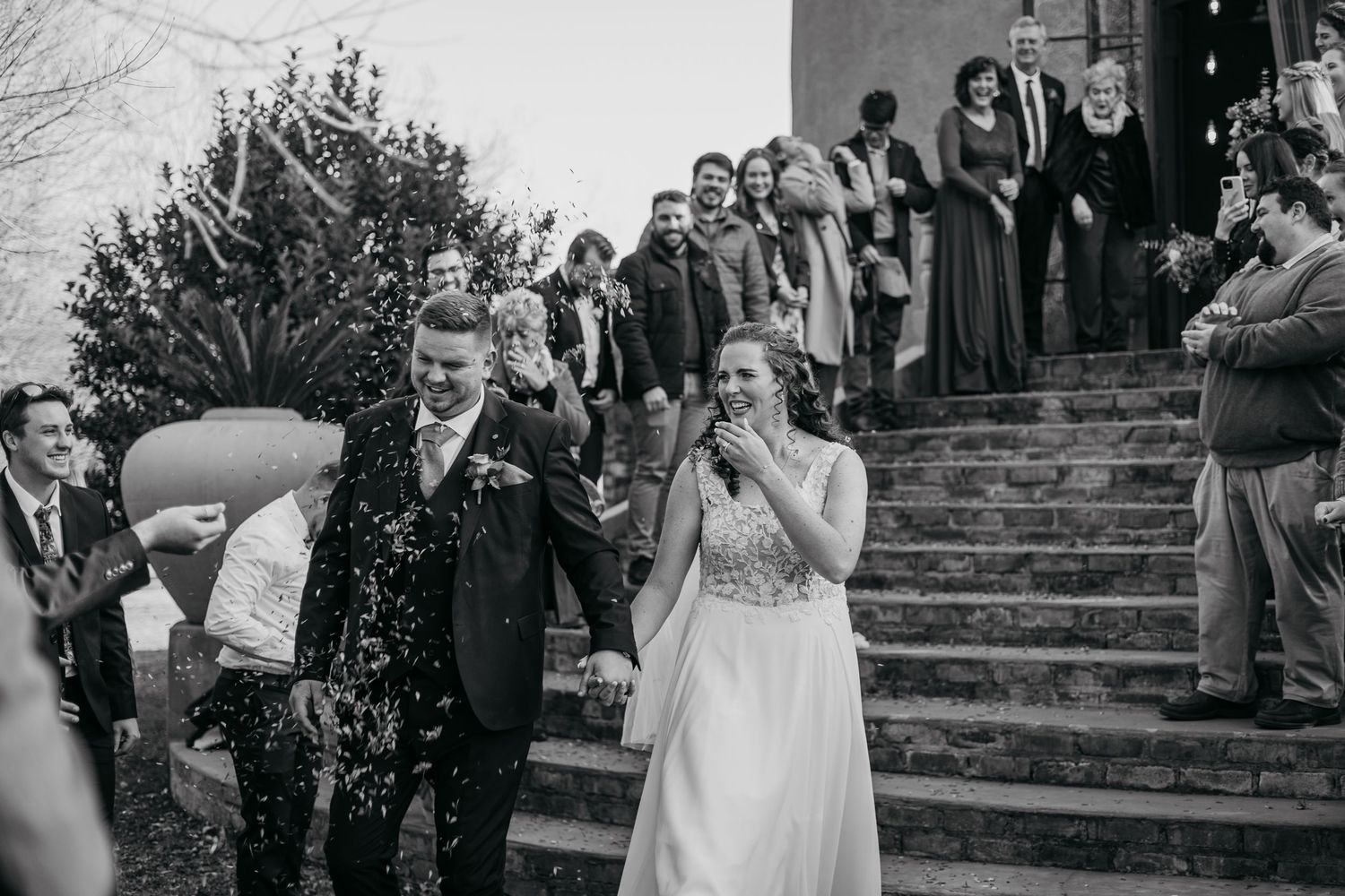 A couple descends stone steps while guests throw confetti at them during a joyful wedding celebration outdoors.