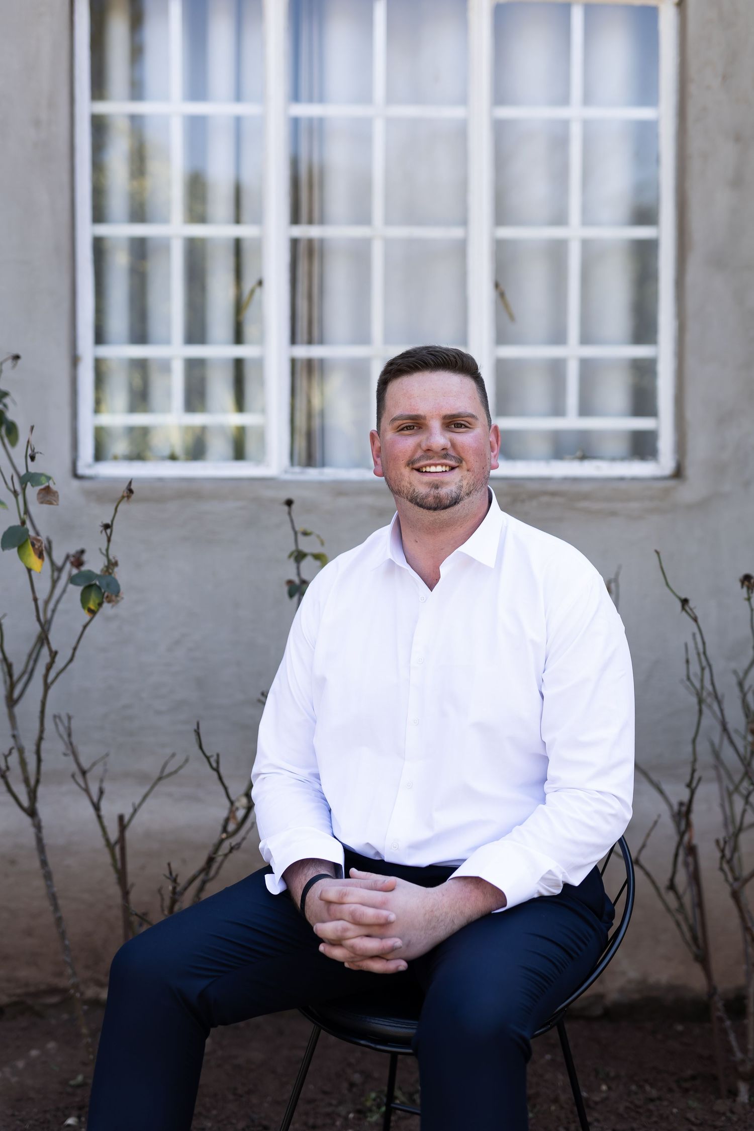 Professional portrait of someone seated outdoors wearing a white dress shirt and dark pants against a gray building.