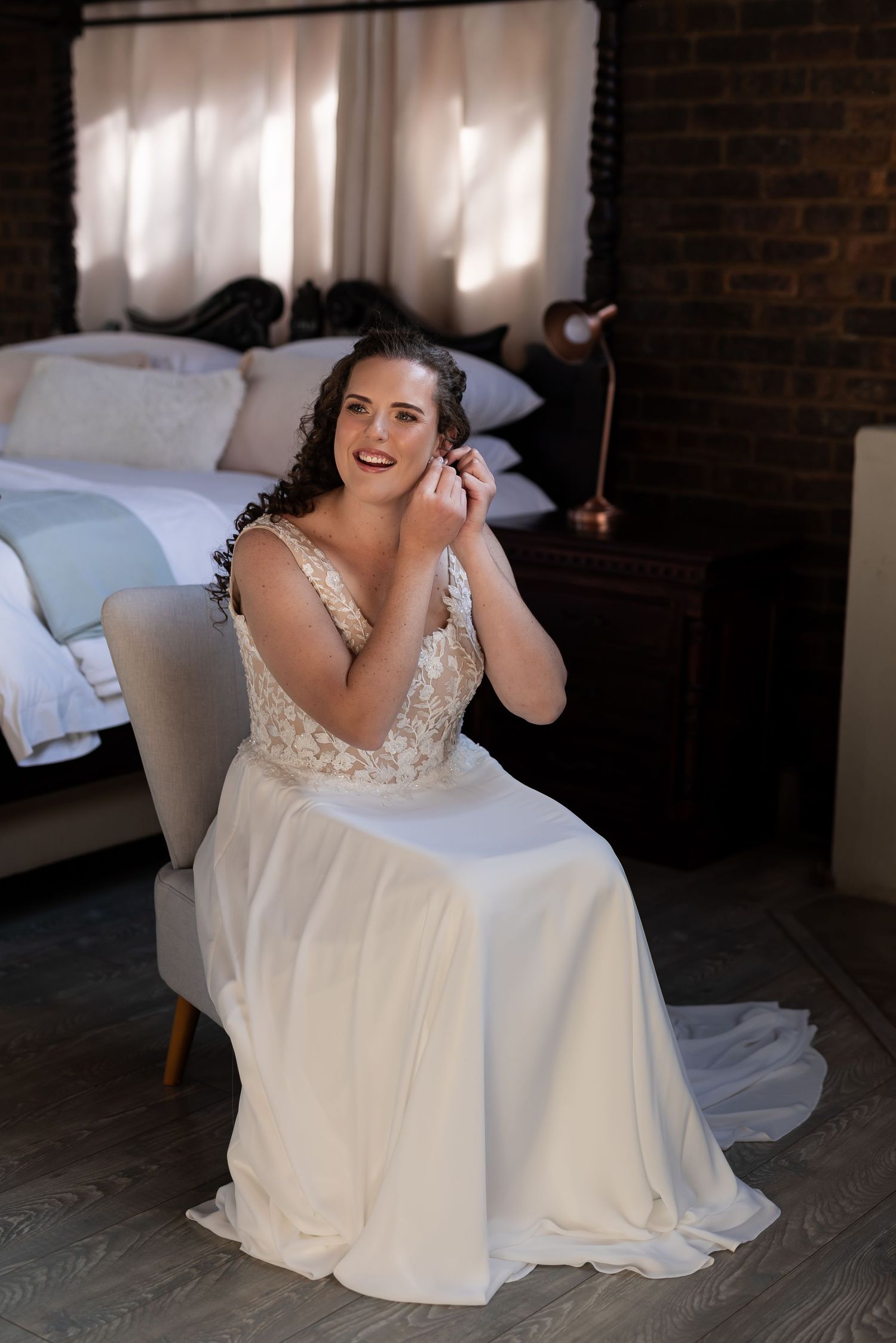 A bride in a flowing white gown with lace detail sits in a grey chair in a bedroom setting.