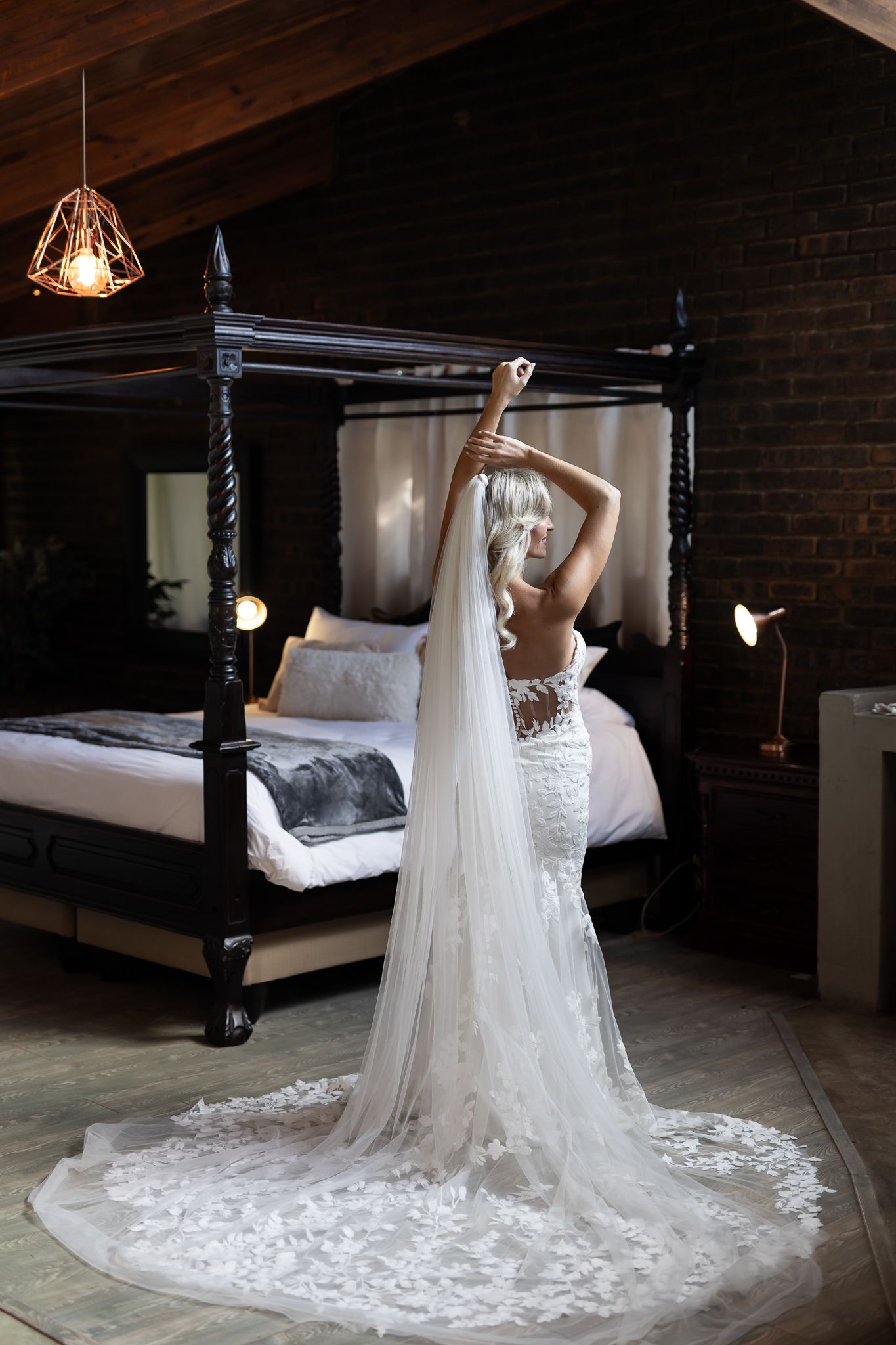 Graceful bridal pose in lace gown with flowing train against dark wooden four-poster bed.