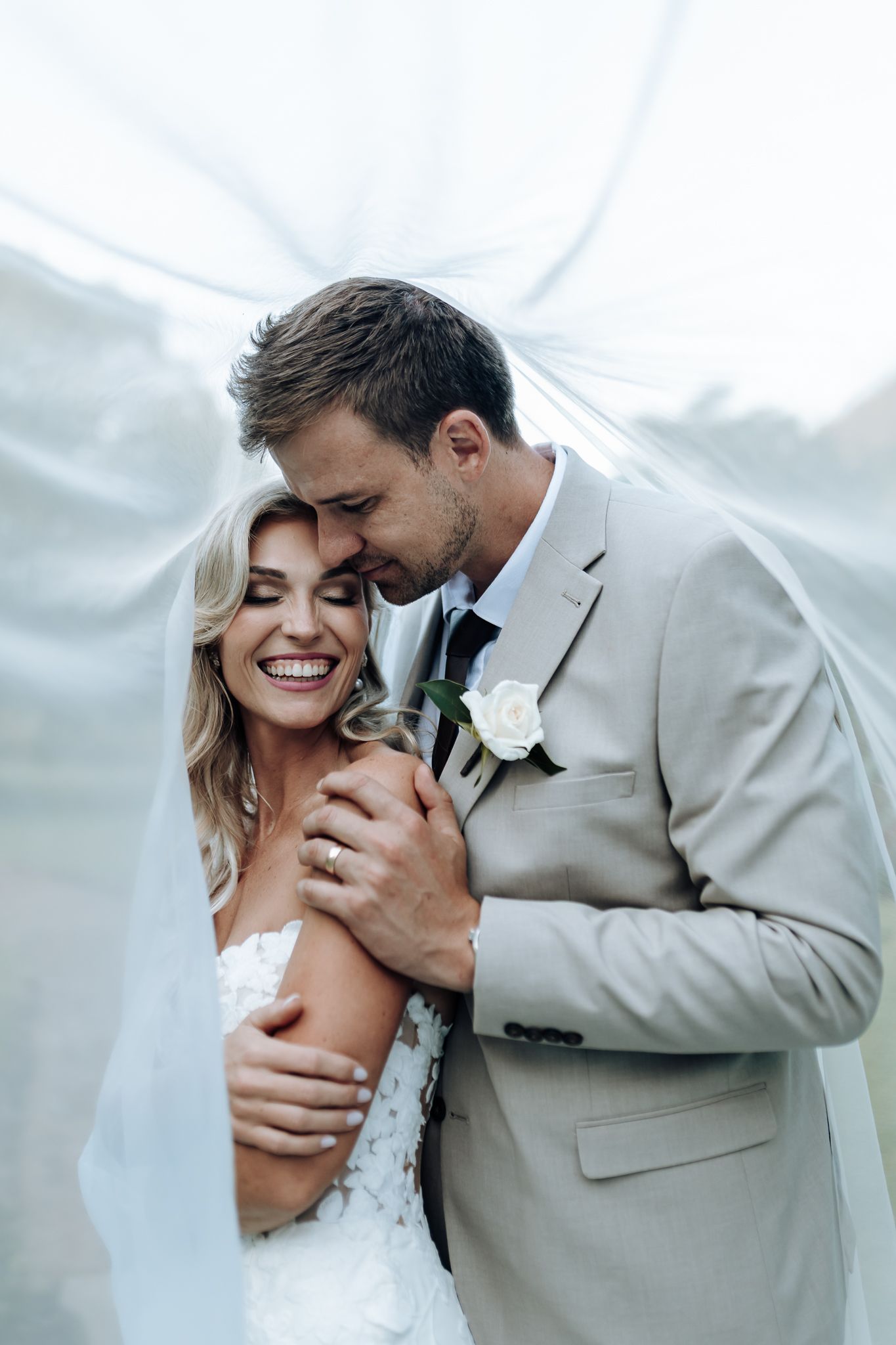 Tender moment between newlyweds under flowing white fabric with soft natural lighting.