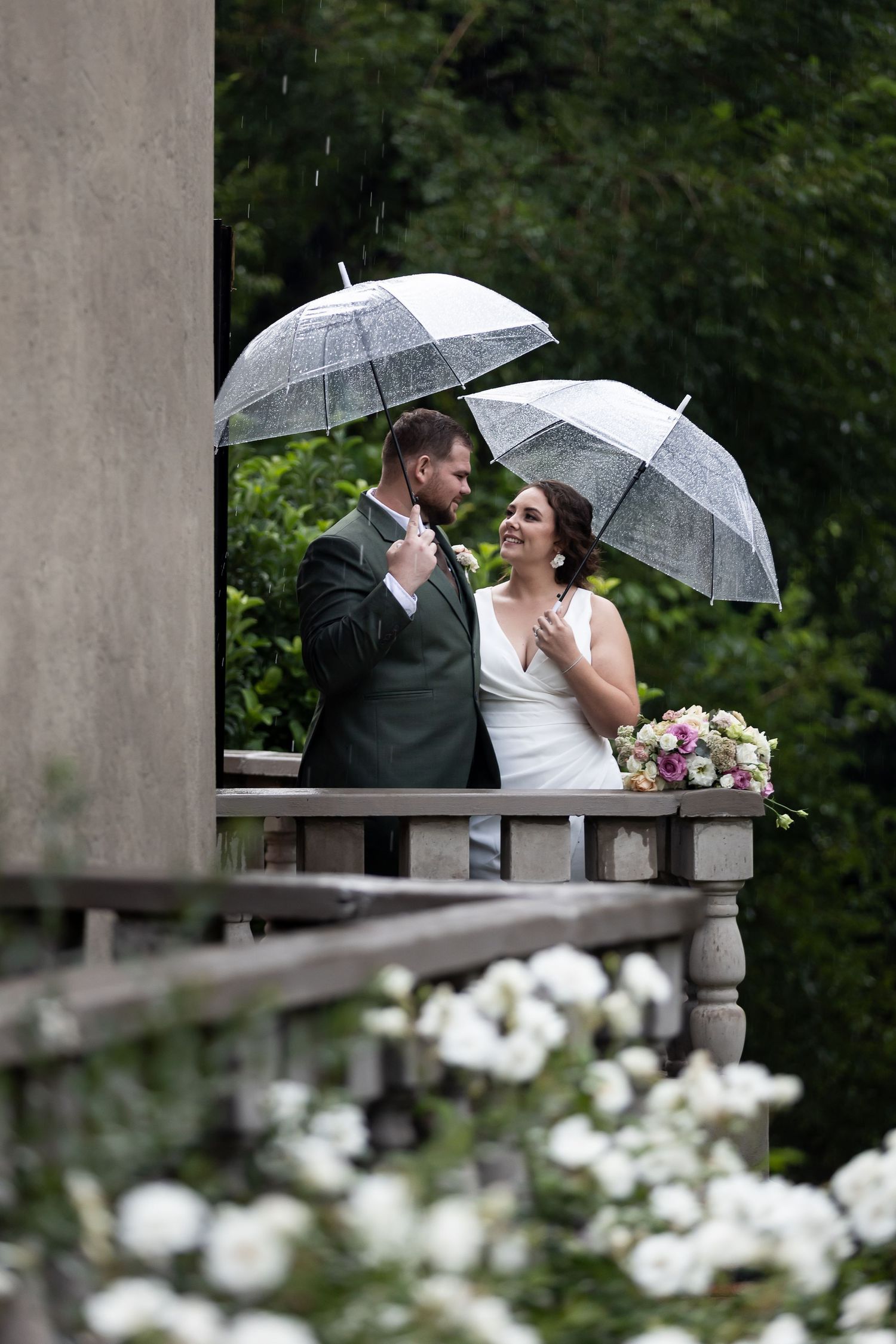 A couple shares an intimate moment under clear umbrellas on a balcony surrounded by white flowers in the rain.