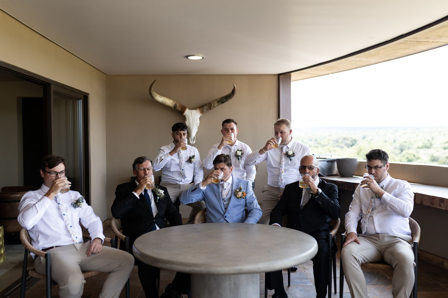 Wedding party in light colored suits sits around circular table on balcony making playful gestures.