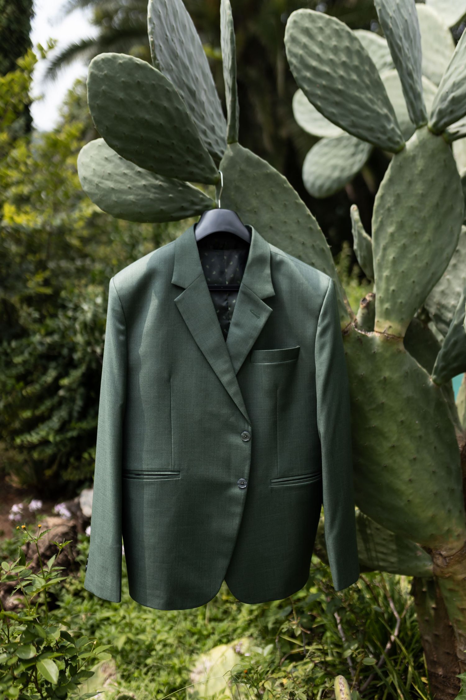Sage green formal suit jacket hanging against large cactus leaves outdoors.