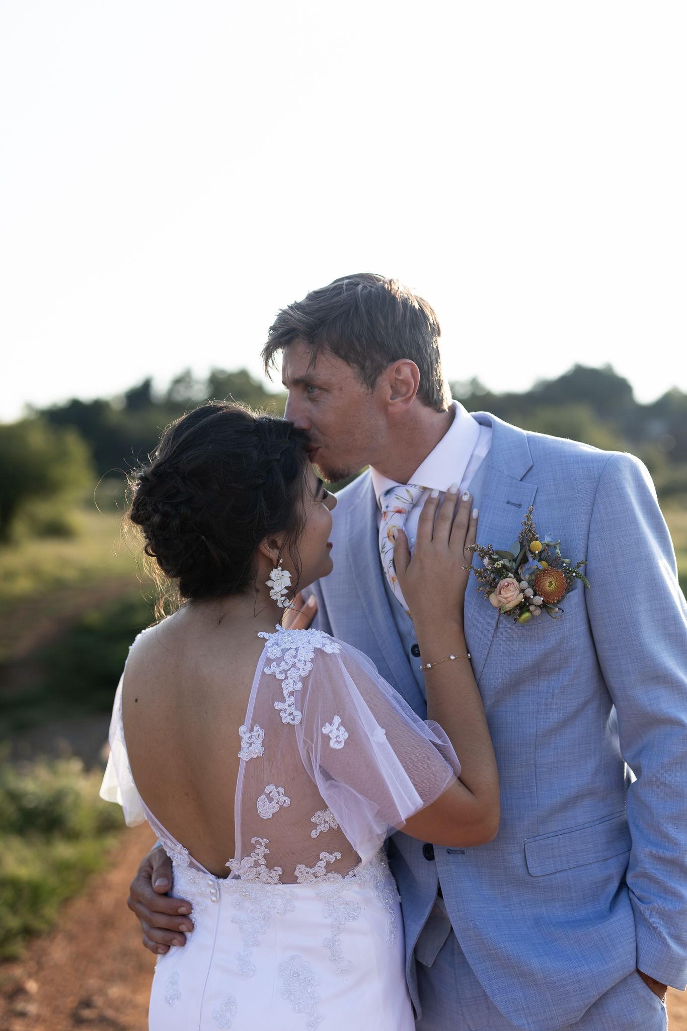 Newlyweds embrace in light blue suit and lace dress against natural outdoor backdrop.
