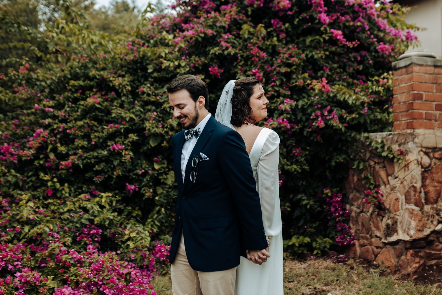 Wedding couple stands back to back against a beautiful flowering bush with pink blooms.