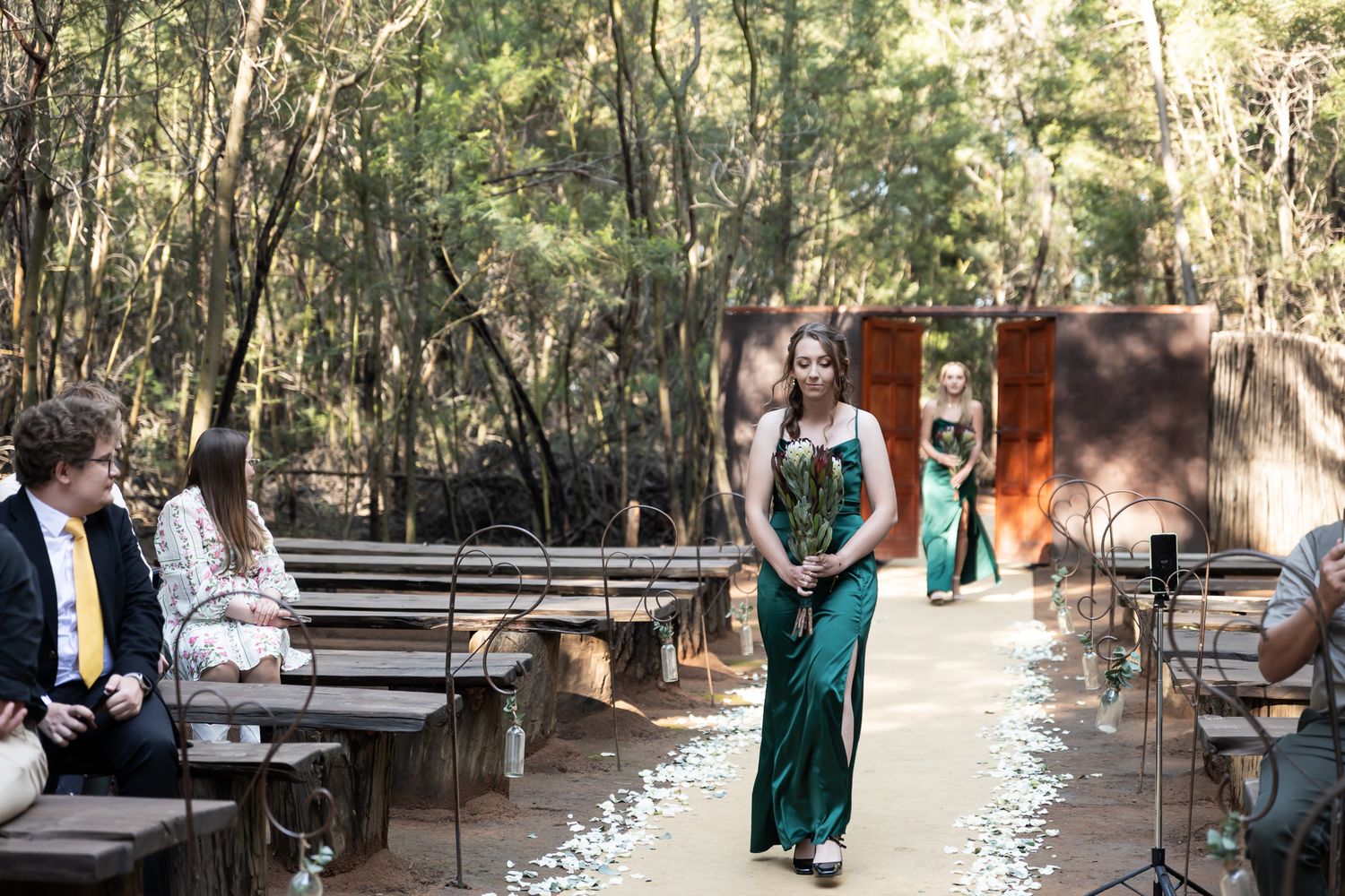 Forest wedding ceremony with emerald-dressed bridesmaid walking past wooden benches.