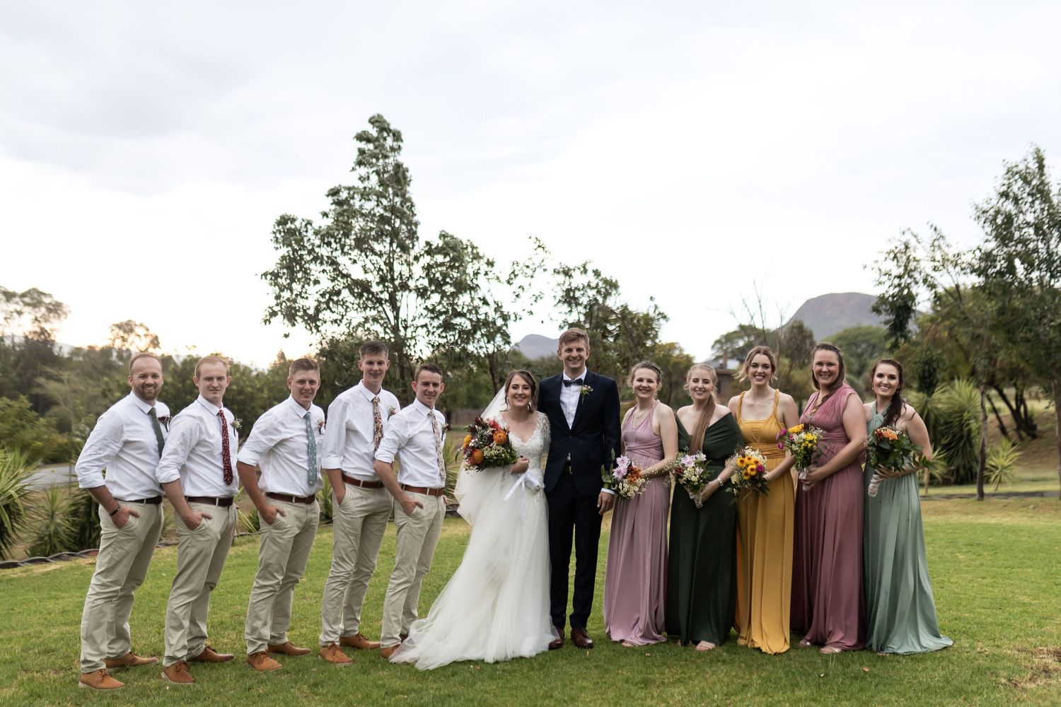 Wedding party in pastel dresses and khaki pants poses on green lawn.