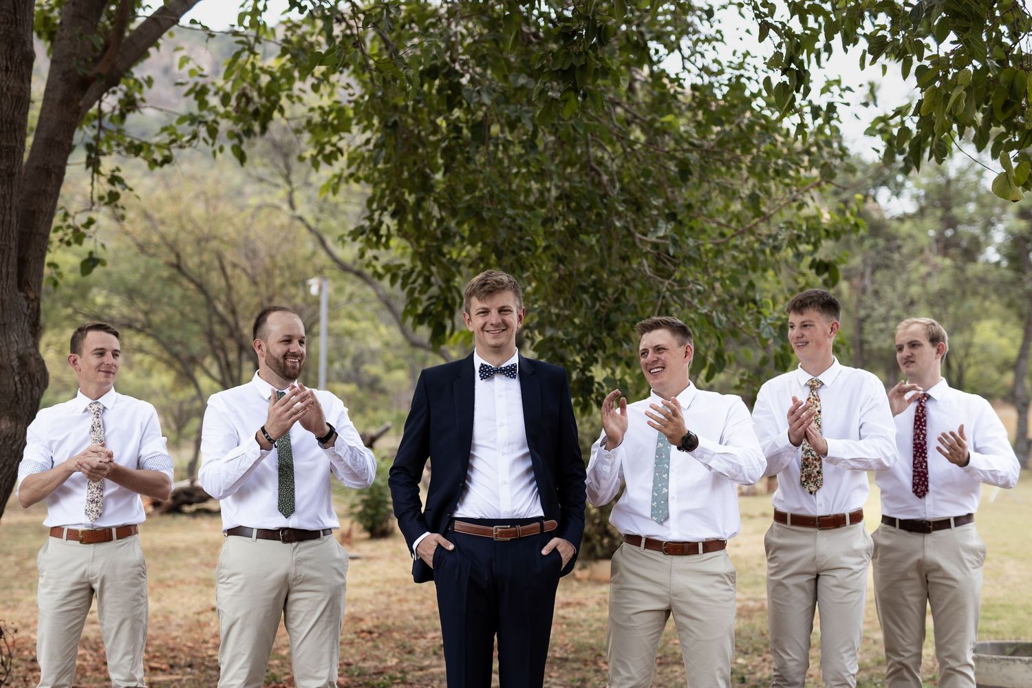 Wedding party poses casually under a tree with the groom in navy surrounded by groomsmen in khakis.