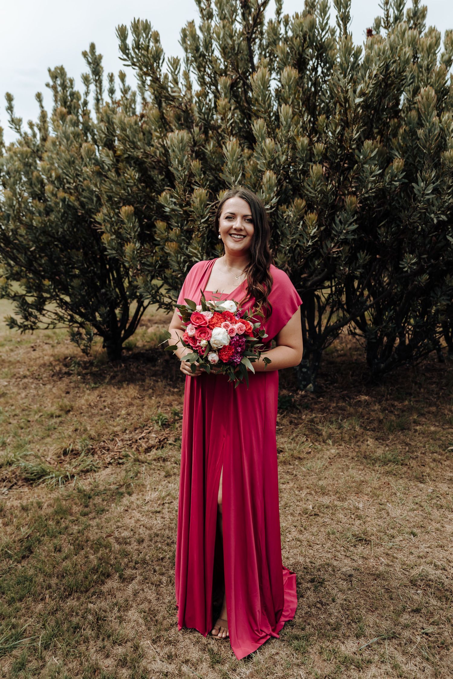 Bridesmaid in flowing red halter dress poses with bouquet against evergreen background.