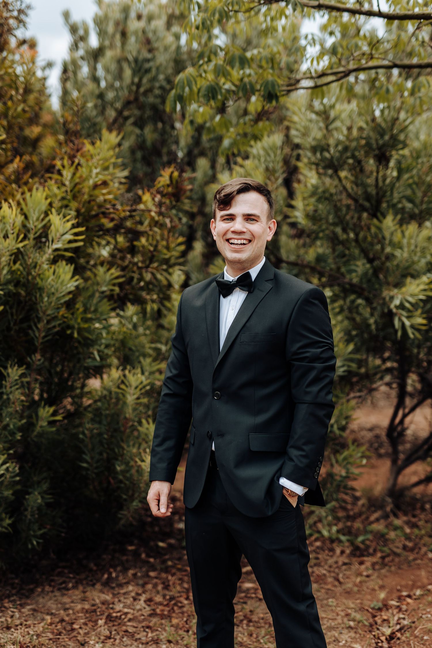 A person in formal black tuxedo and bowtie smiles warmly while standing in a natural outdoor setting.