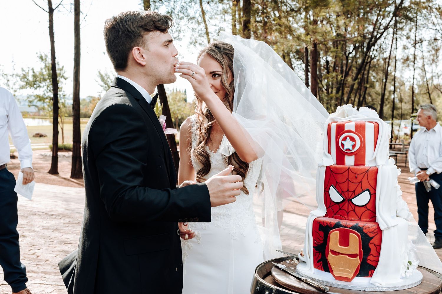 Wedding couple feeds each other cake beside a creative superhero themed dessert display.