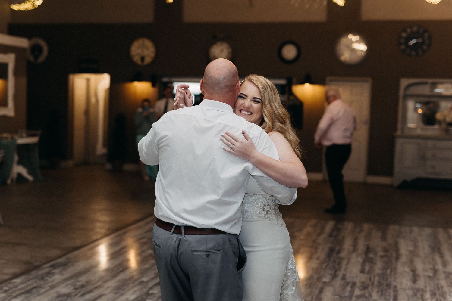 Romantic slow dance moment on wooden floor with string lights and decor in background.
