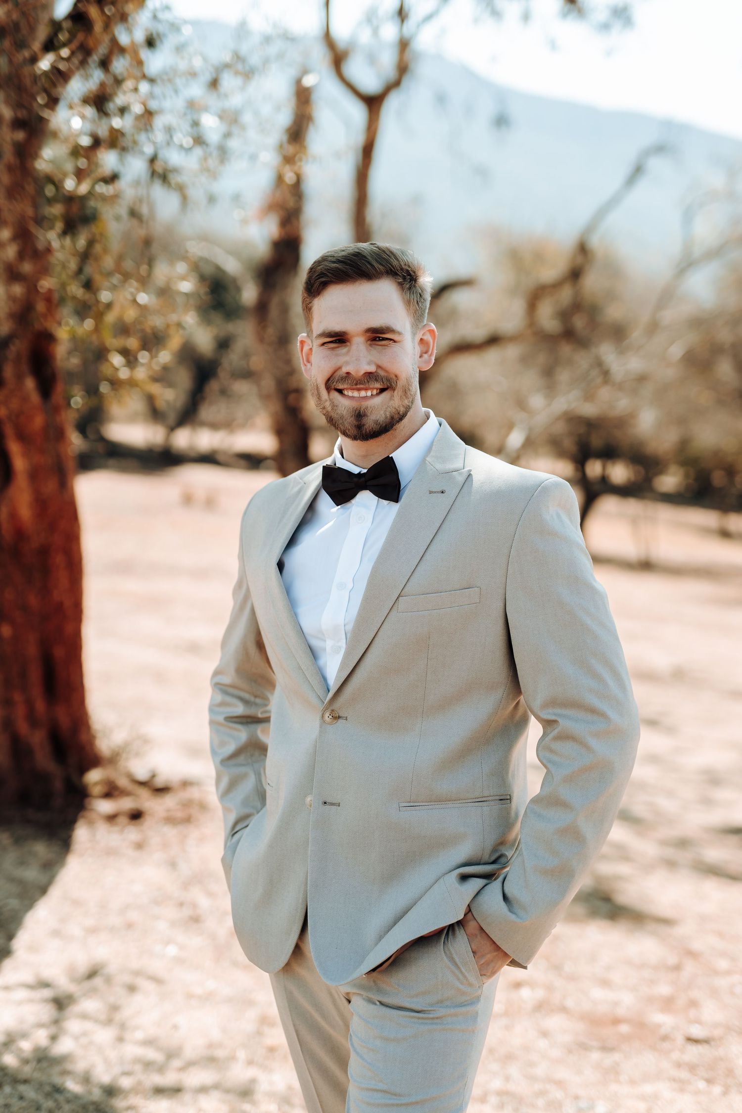 Wedding portrait of groom in light beige suit and bow tie against desert landscape.