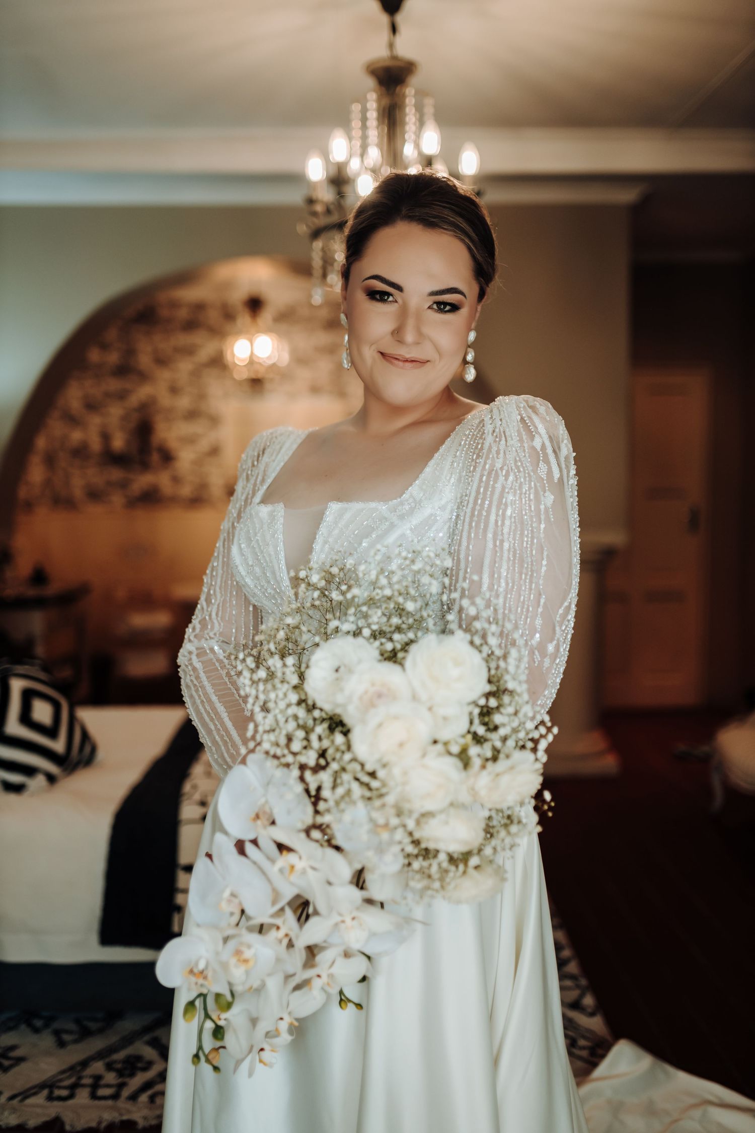 Classic bridal pose showing detailed wedding dress with beaded sleeves and white orchid bouquet in elegant room setting.