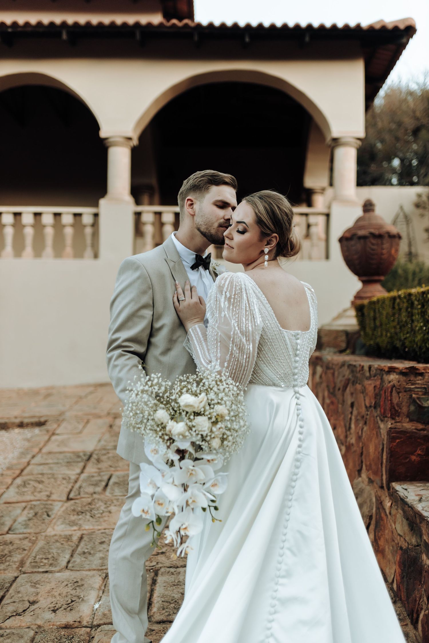 Intimate wedding moment captured against cream-colored arched colonnade and stone patio.