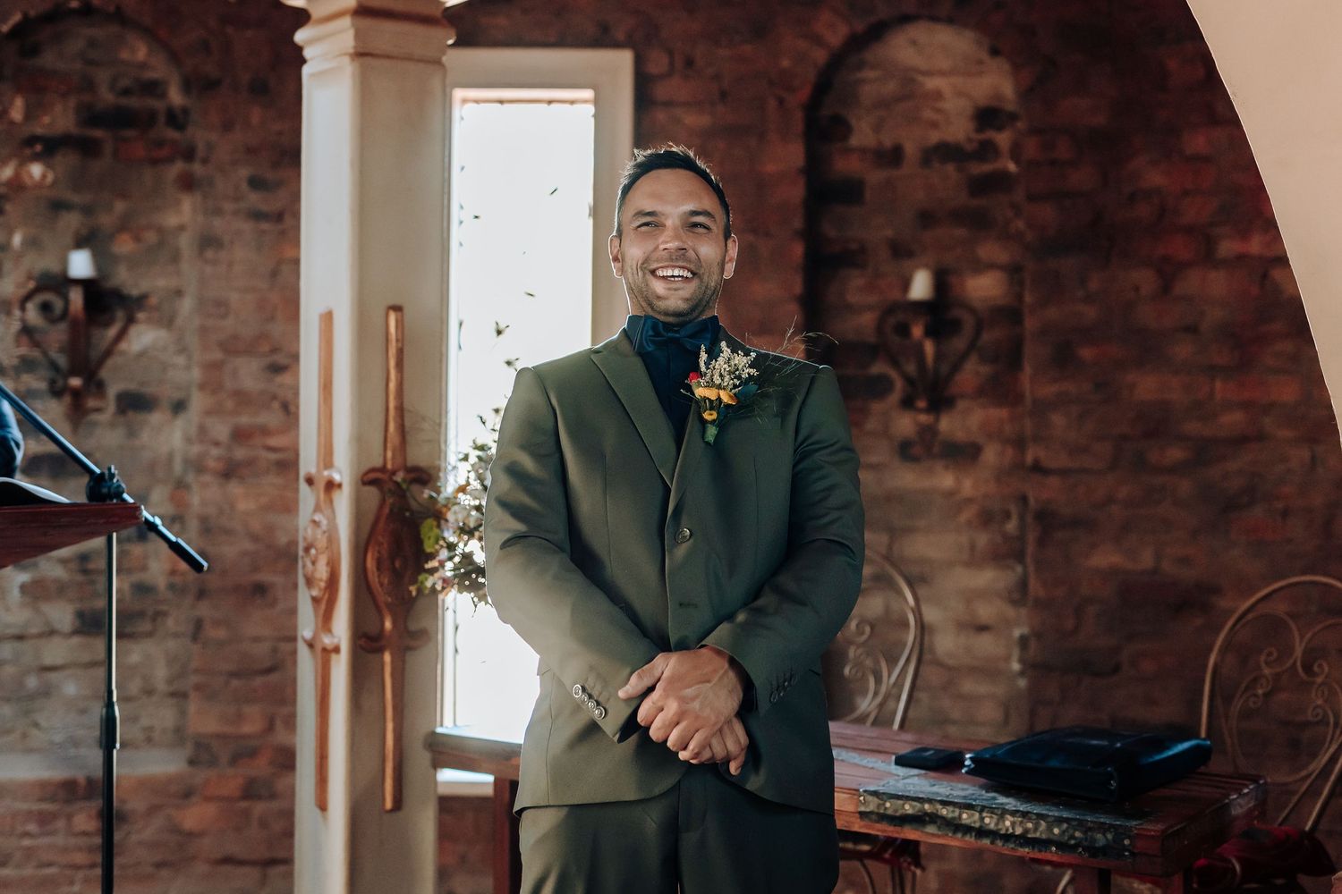 Person in olive green suit with boutonniere smiling at wedding venue.