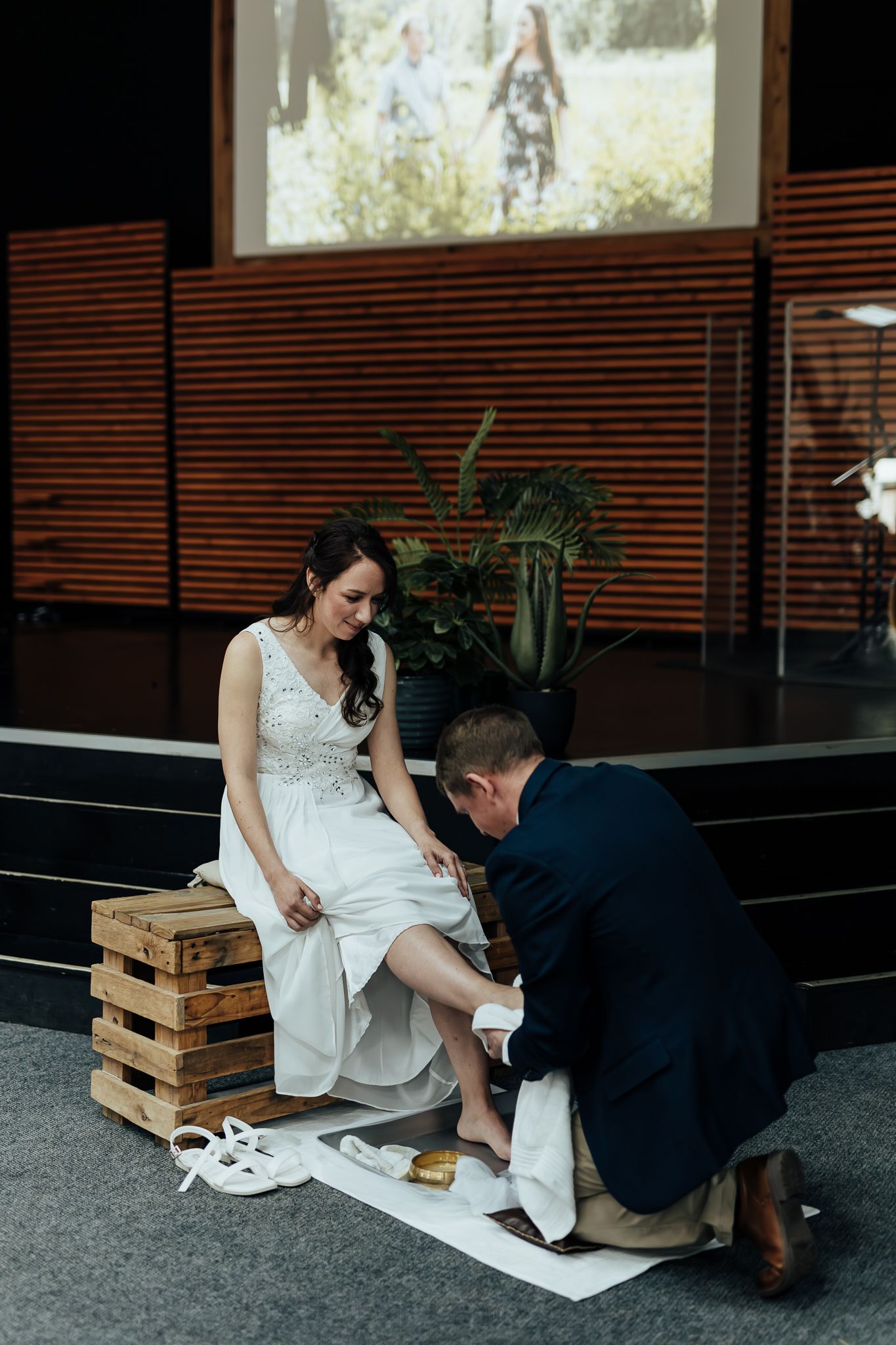 A sweet wedding moment as groom helps put on his bride's shoes while she sits on wooden crates.