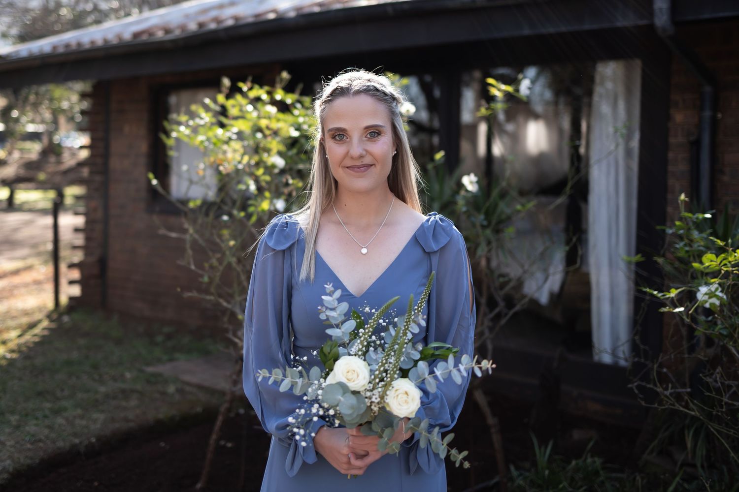 Person in dusty blue dress holding white rose bouquet with eucalyptus outside rustic building.