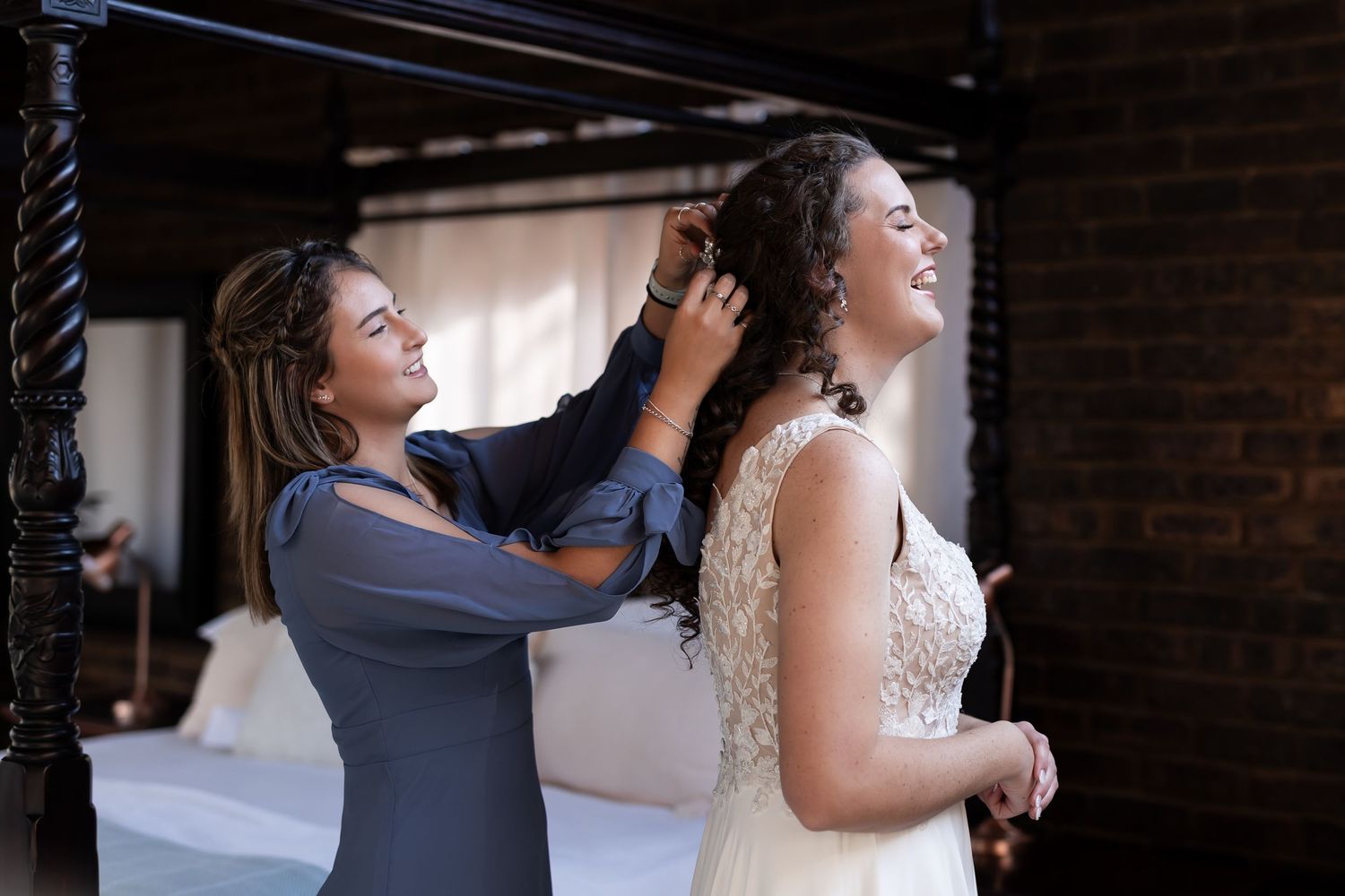 Bride in a white lace gown laughs while getting her hair styled in a rustic brick venue.