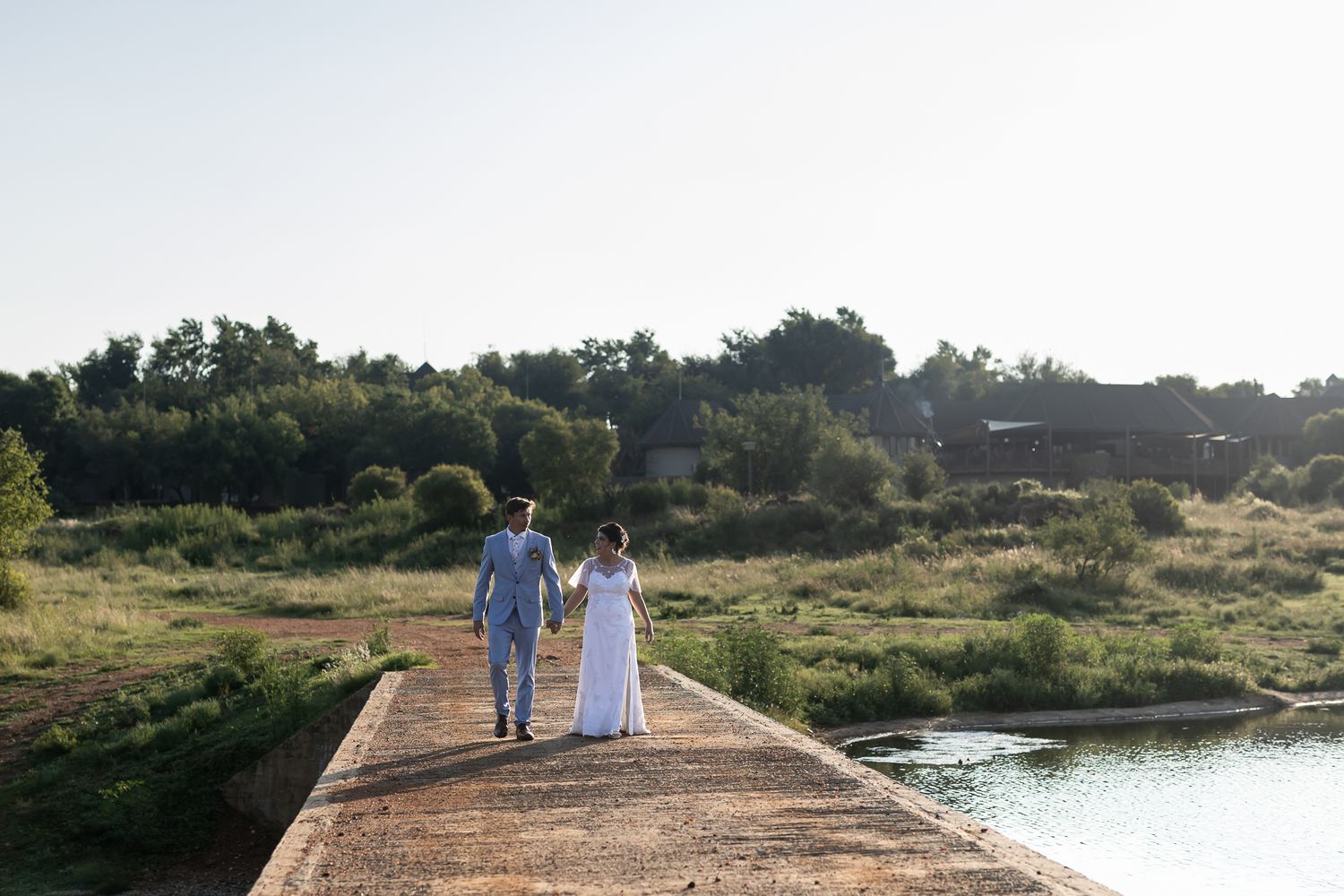 Newlyweds walking hand in hand on concrete pier by waterfront at sunset.