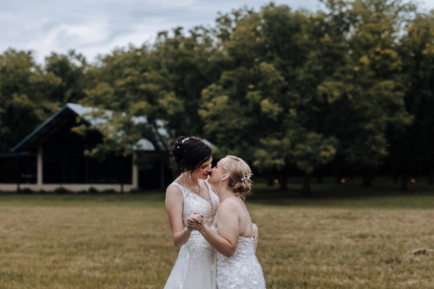 Two people in white wedding dresses standing together in a field against a cloudy sky.