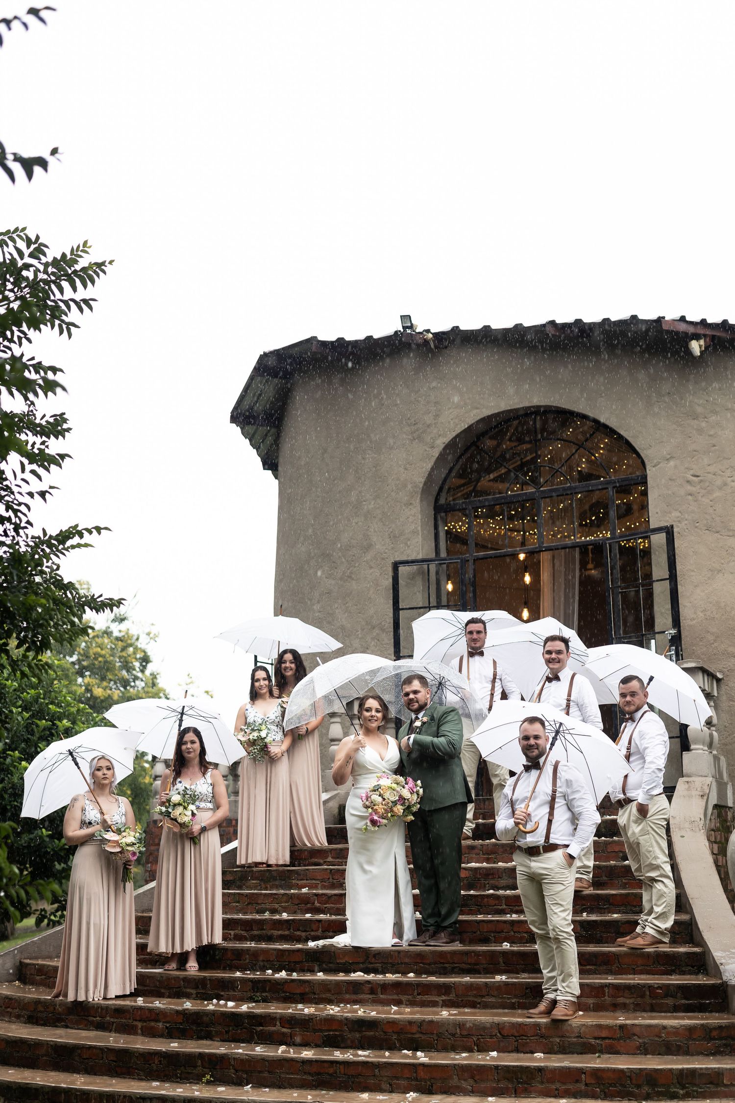 Wedding party arranged on stone steps holding white umbrellas in front of arched doorway.