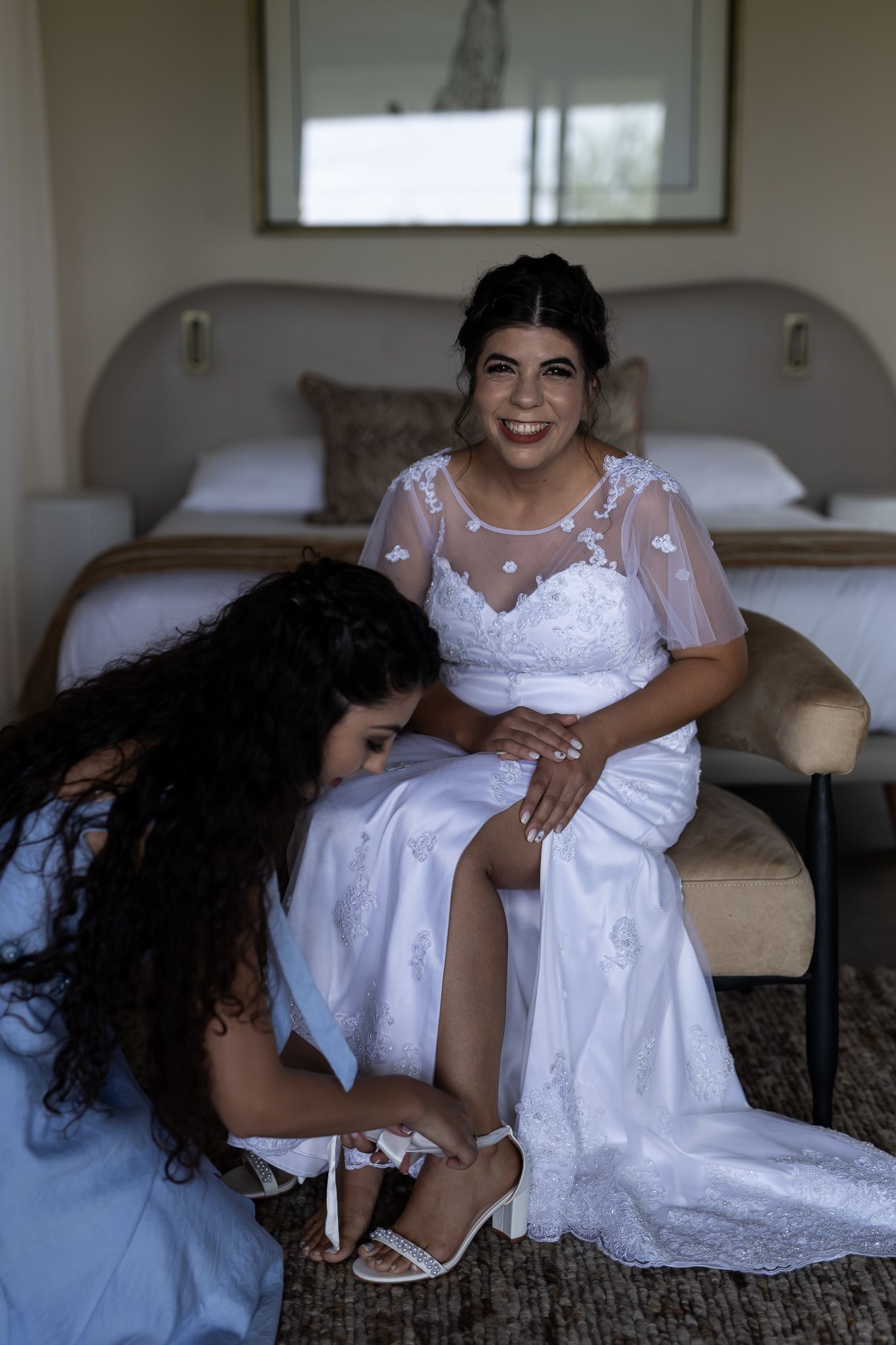 Someone in a wedding dress sits on a bed while getting their shoes adjusted.