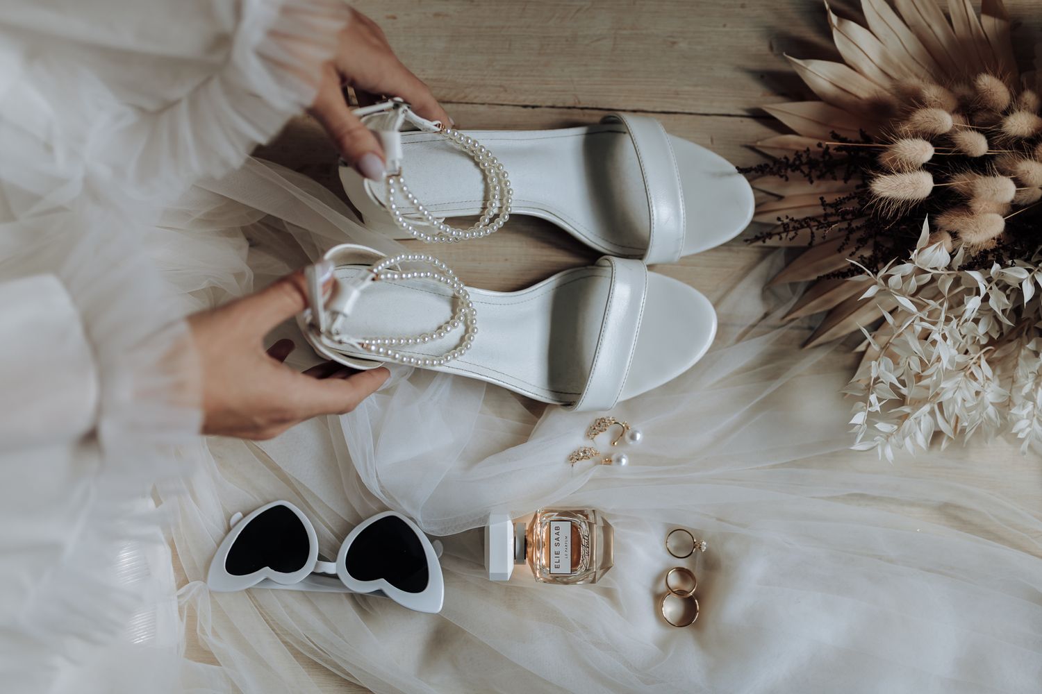 Elegant bridal accessories including white sandals, sunglasses, watch and jewelry laid on white fabric.