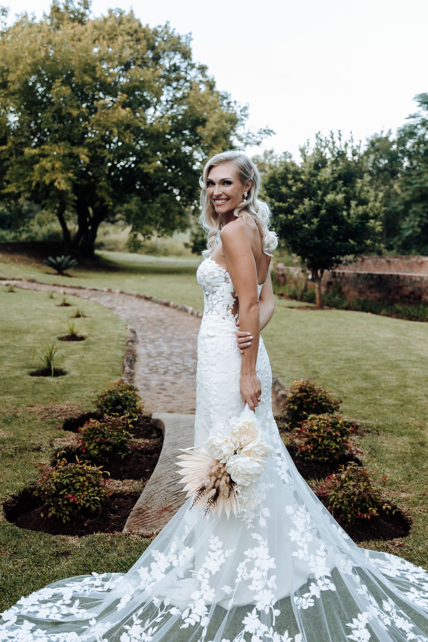 Elegant bride wearing a fitted lace gown with a dramatic train poses in a landscaped garden.
