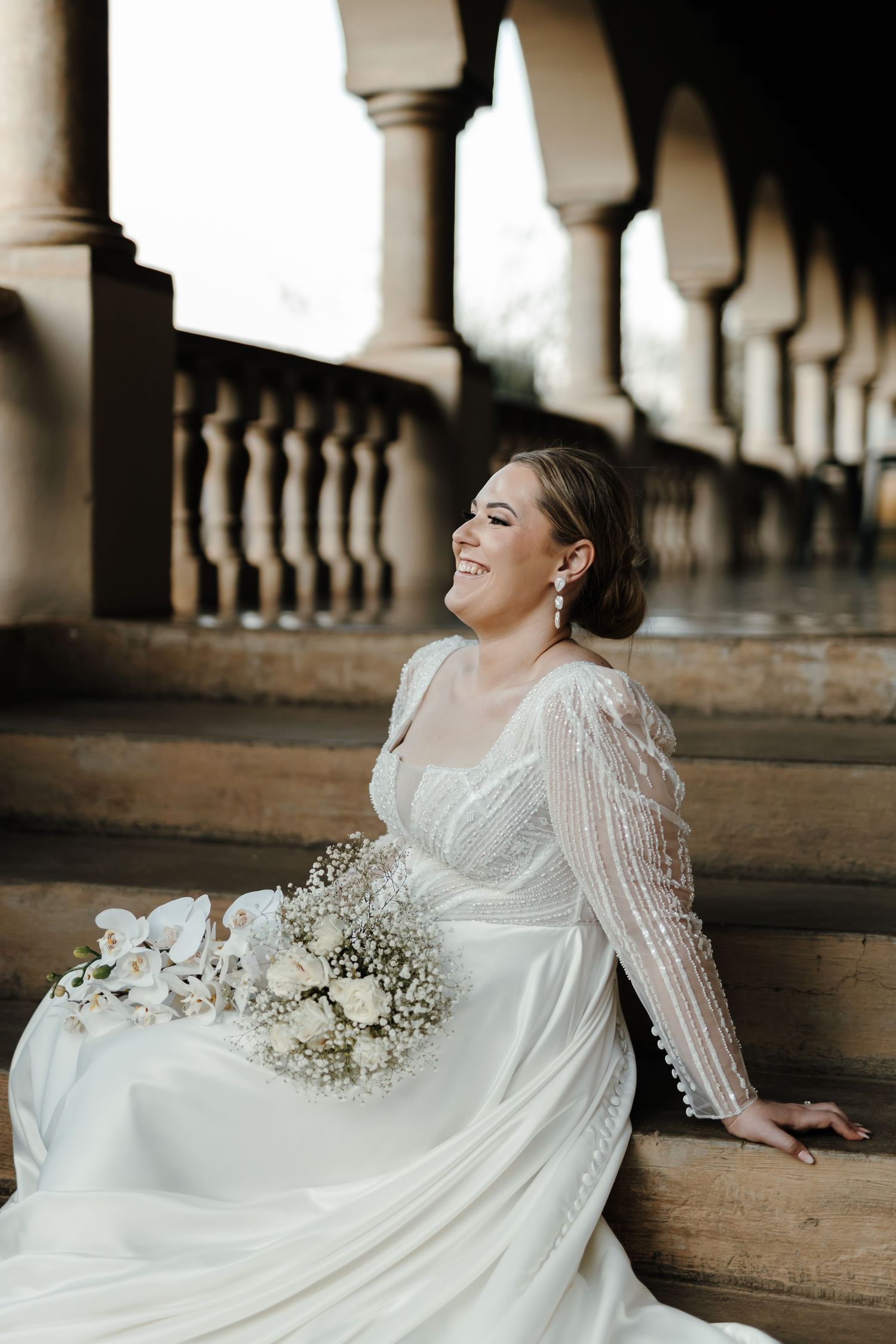 Joyful bride in lace sleeve wedding gown sitting on colonnade steps with white bouquet.