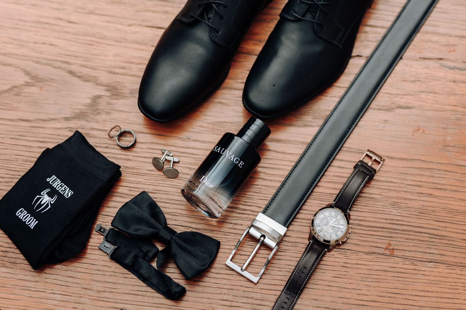 Groom's accessories including black dress shoes, bow tie, belt and watch on wooden surface.