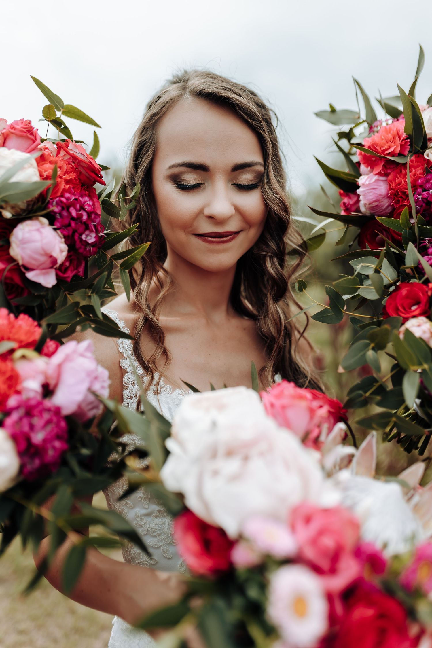 Bride in white dress surrounded by pink and red flowers with eyes closed in serene outdoor portrait.