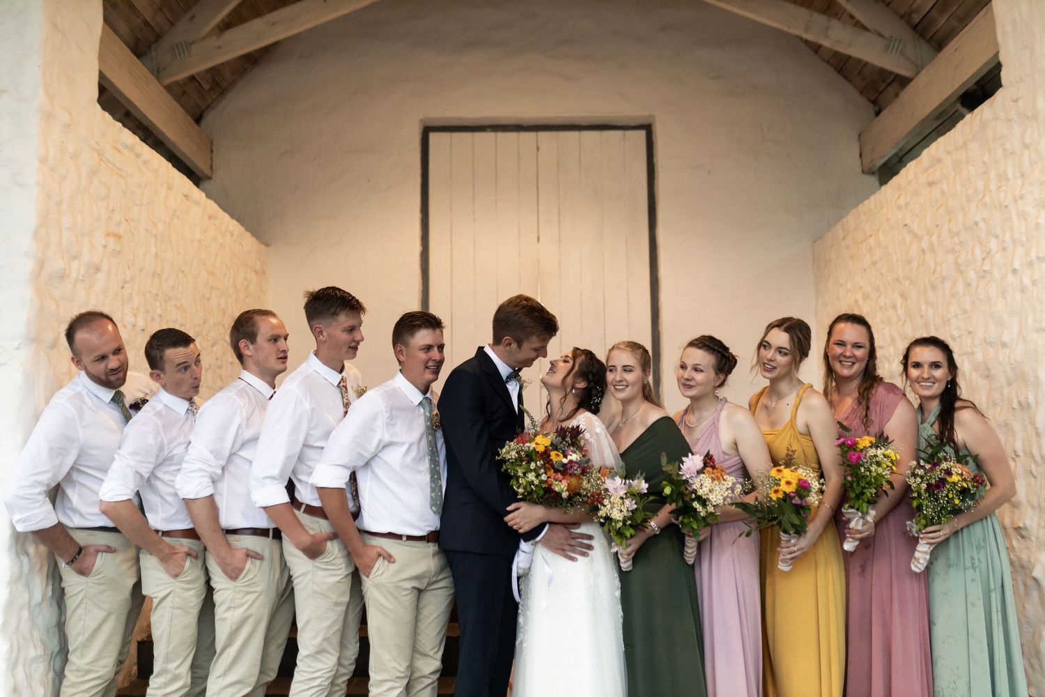 Wedding party poses together under an archway with pastel bridesmaid dresses and khaki groomsmen attire.