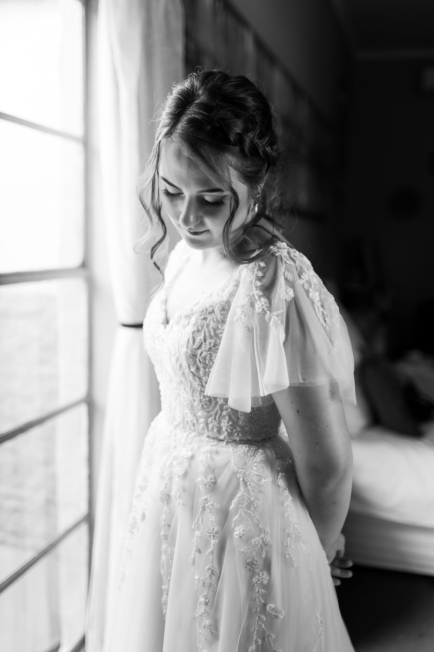 An elegant wedding dress with delicate lace details and flutter sleeves captured in dramatic window light.