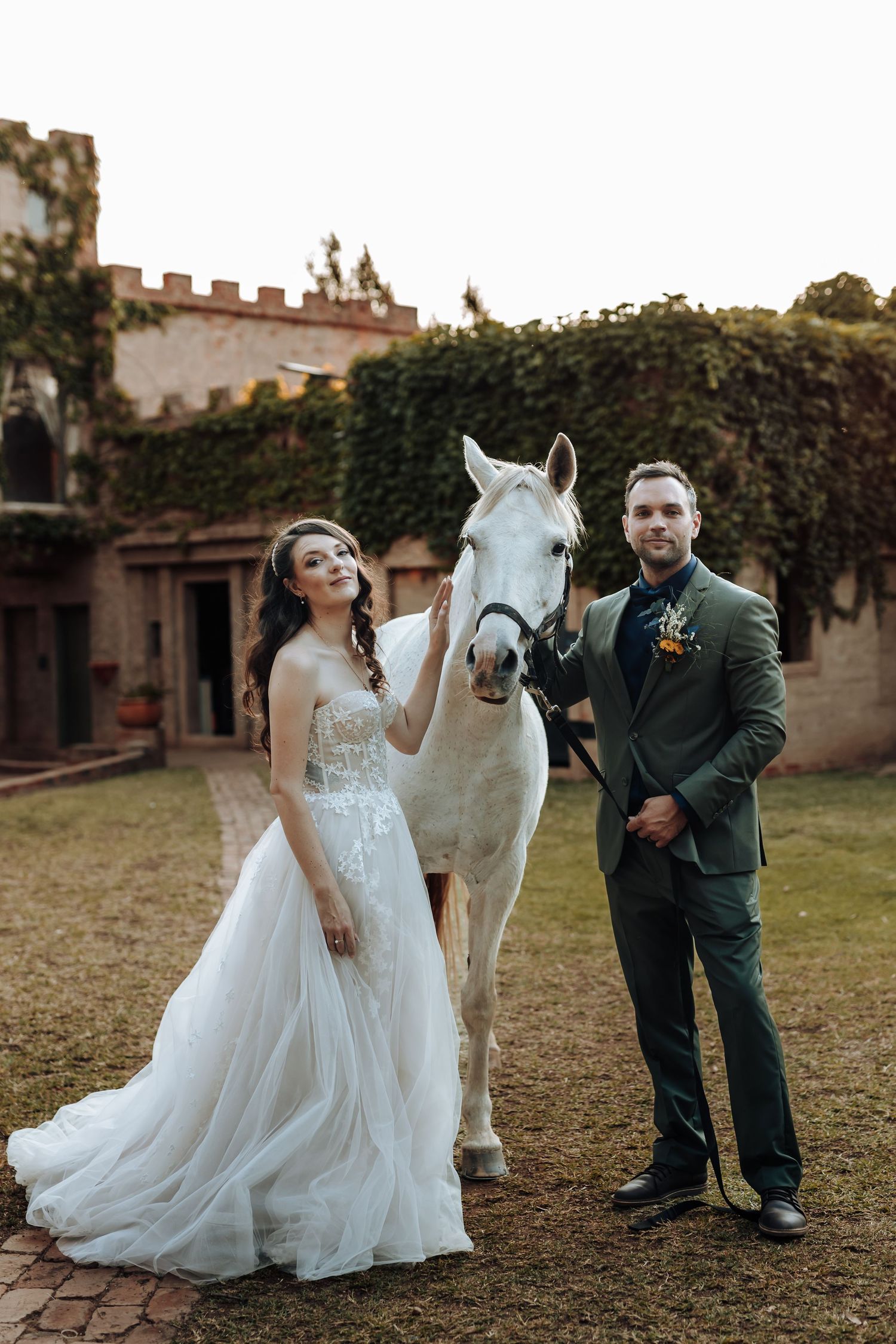 Wedding portrait featuring couple and white horse against rustic castle architecture.