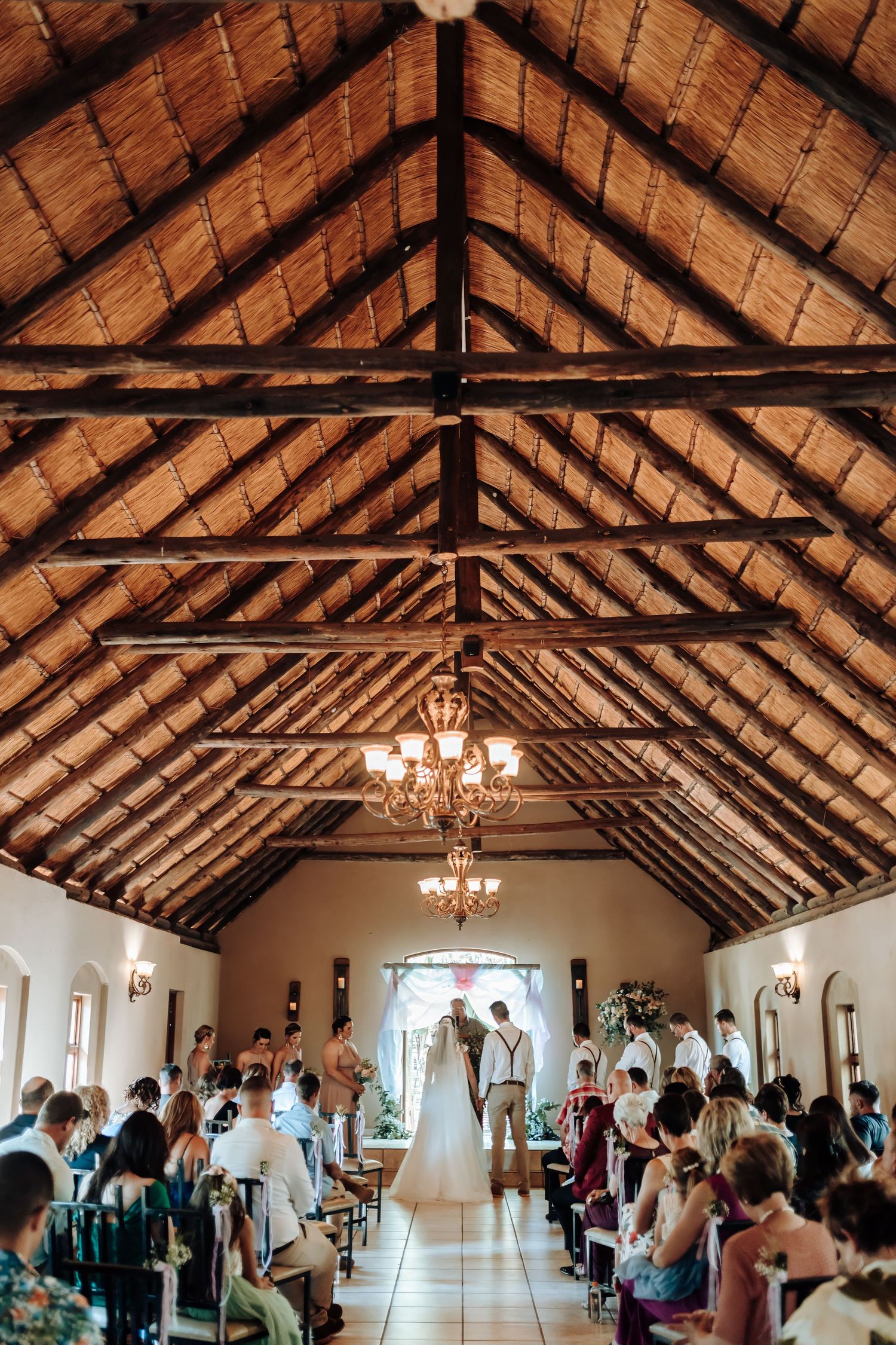 Wedding ceremony in beautiful chapel with exposed wooden beams and elegant chandeliers overhead.