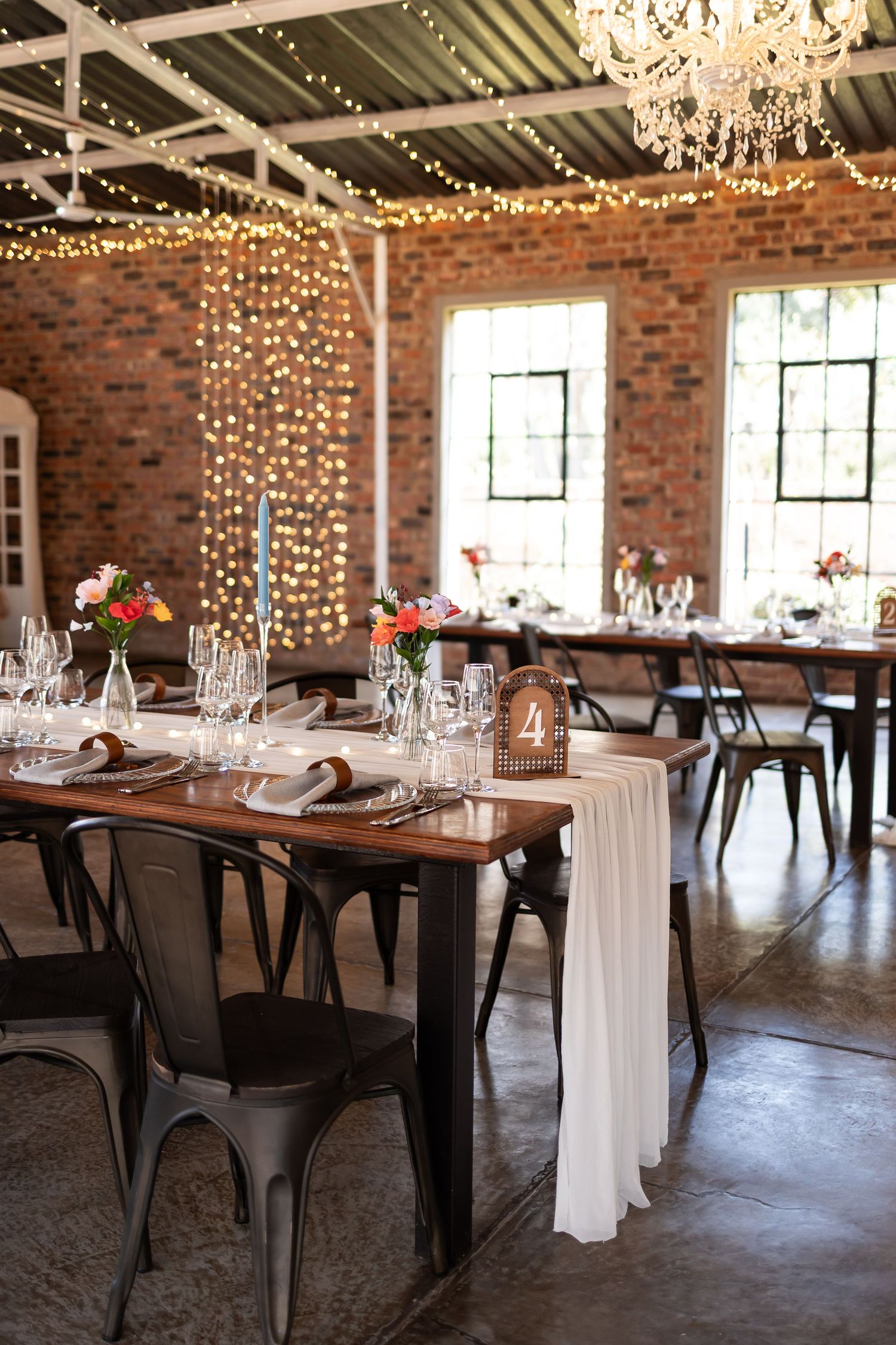 Rustic industrial wedding reception venue with exposed brick walls, string lights, chandeliers and elegant table settings.