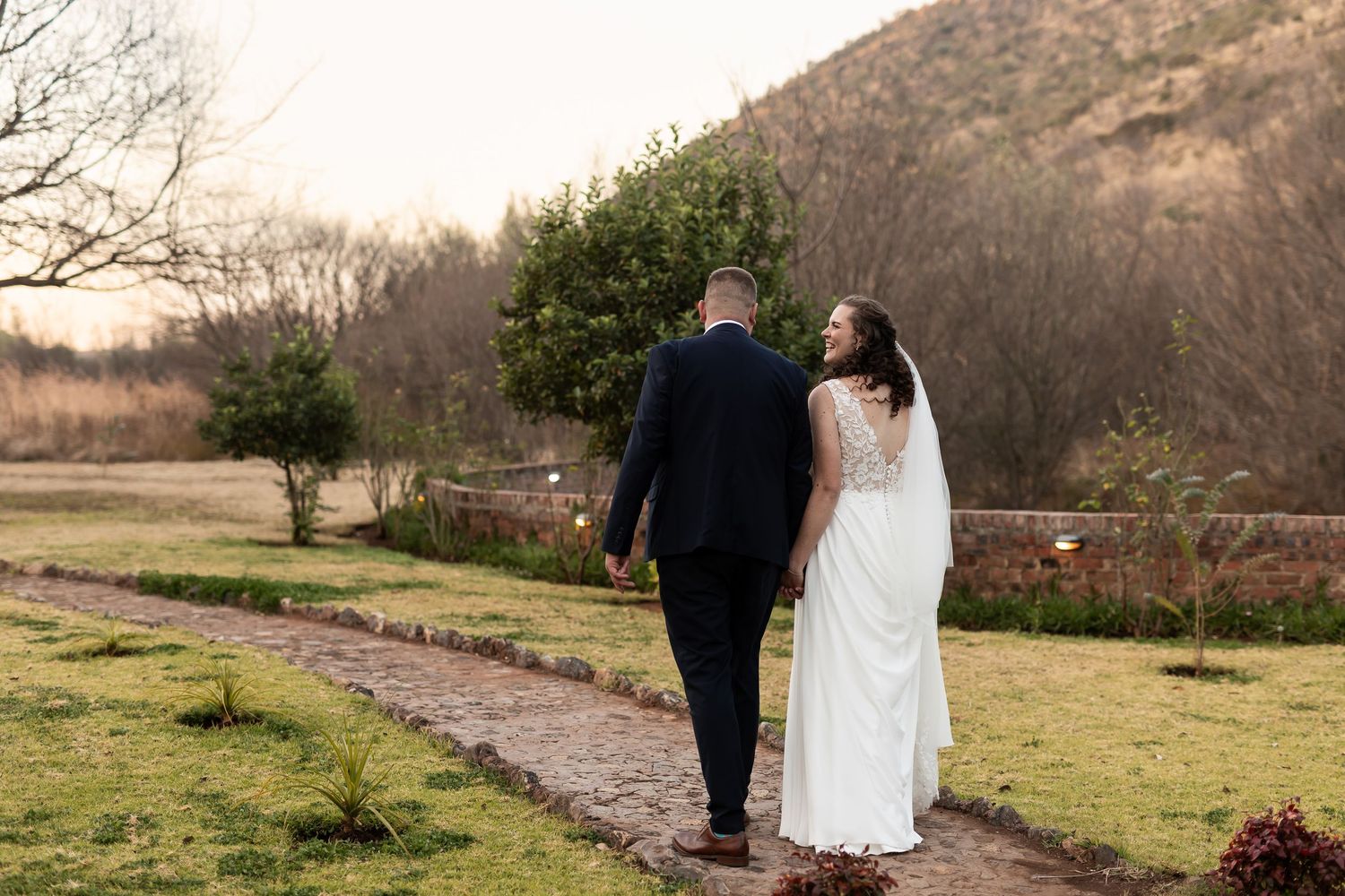 A bride and groom walk hand in hand down a stone path surrounded by nature on their wedding day.