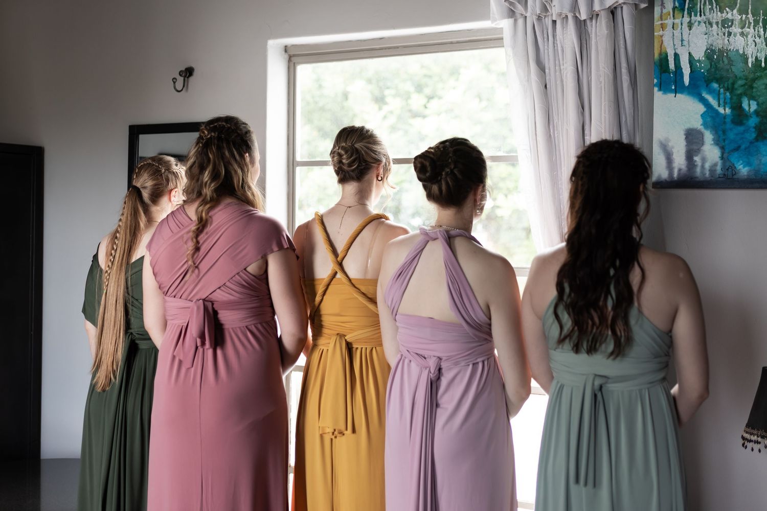 Bridesmaids in pastel dresses stand together looking out a window.