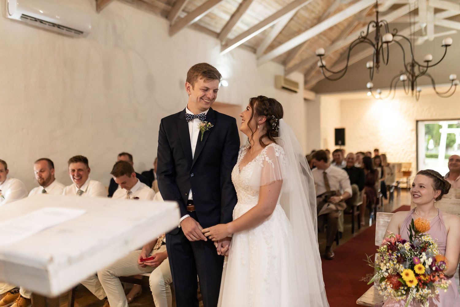 A couple stands before their guests during a wedding ceremony in a rustic indoor venue.