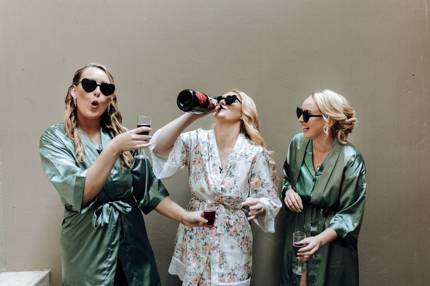 Three friends in robes drink from wine bottles while posing against a wall.