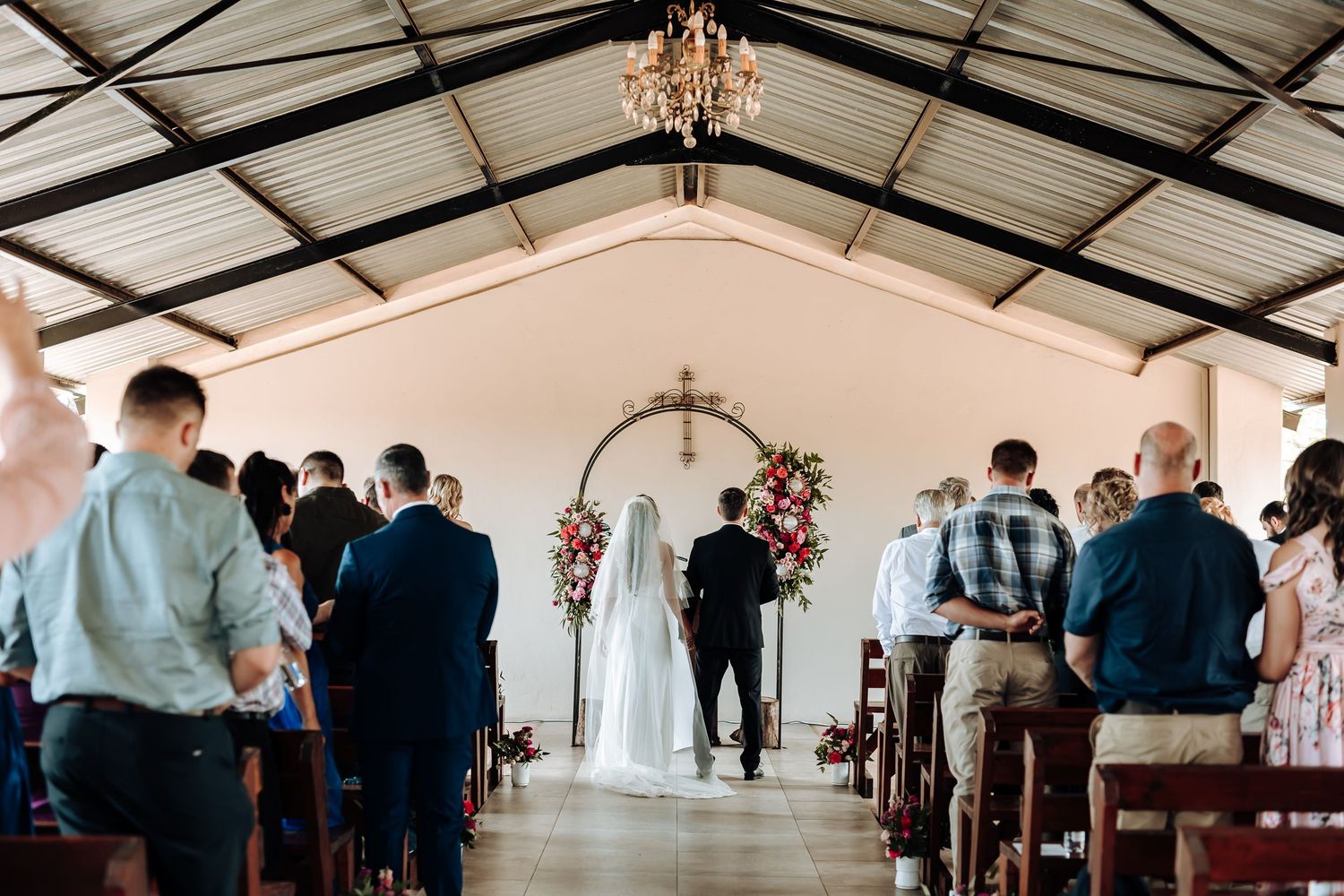 Wide view of wedding ceremony in covered venue with floral arch and chandelier.
