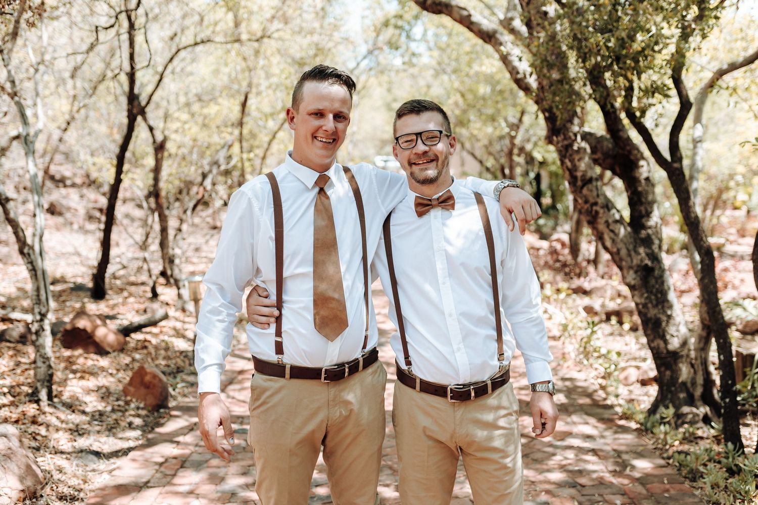 Wedding party members wearing brown suspenders and ties pose casually in a rustic outdoor setting.